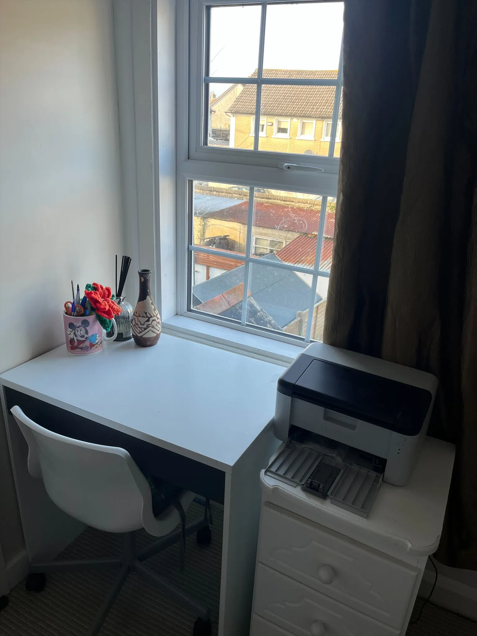 Small workspace with a white desk and chair by a multi-pane window, desk items (cups, vase) on the left and a printer on the right, dark curtain partially drawn.