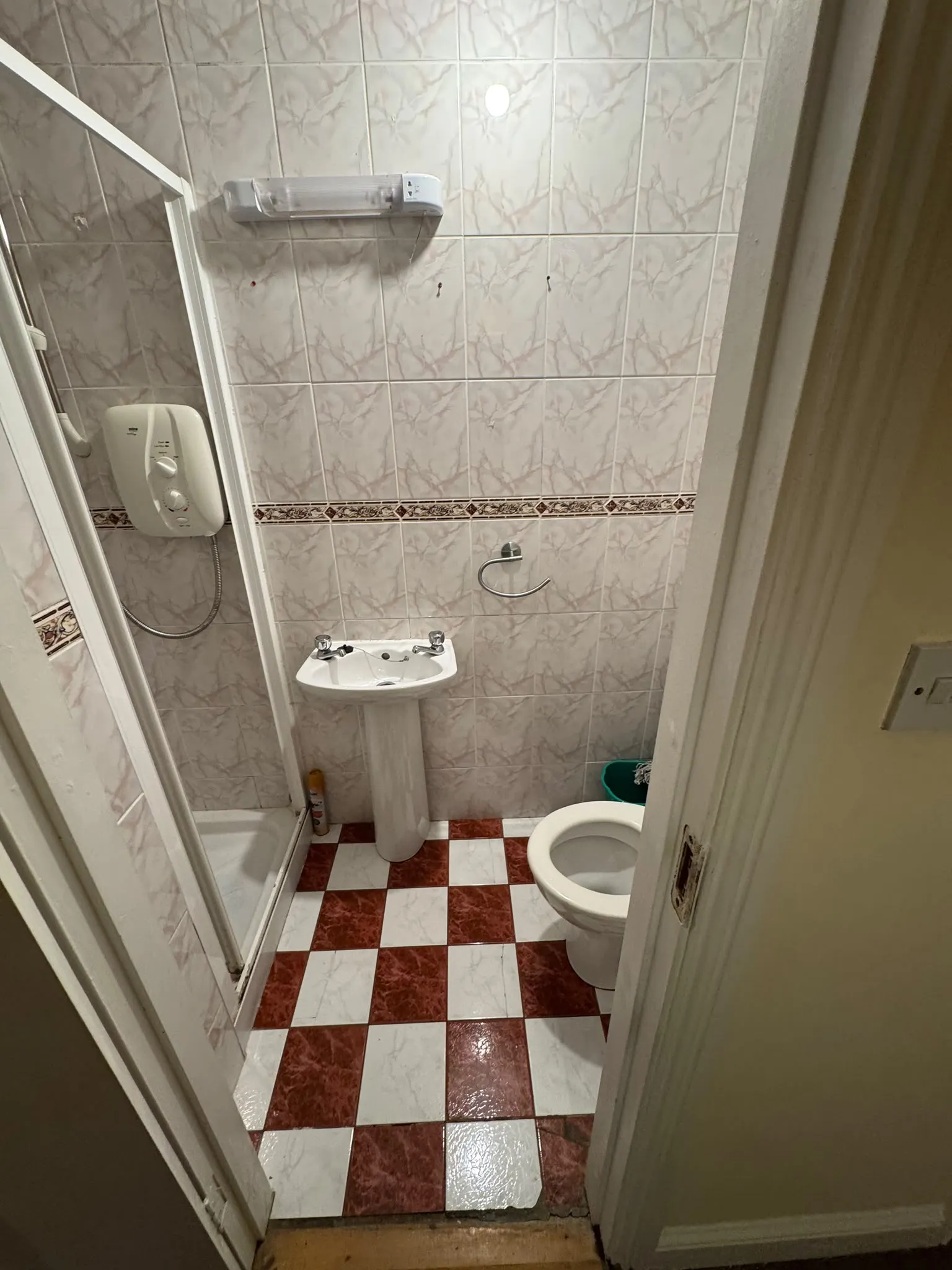 Small bathroom with a pedestal sink, toilet, corner shower and checkered red-and-white tiled floor.