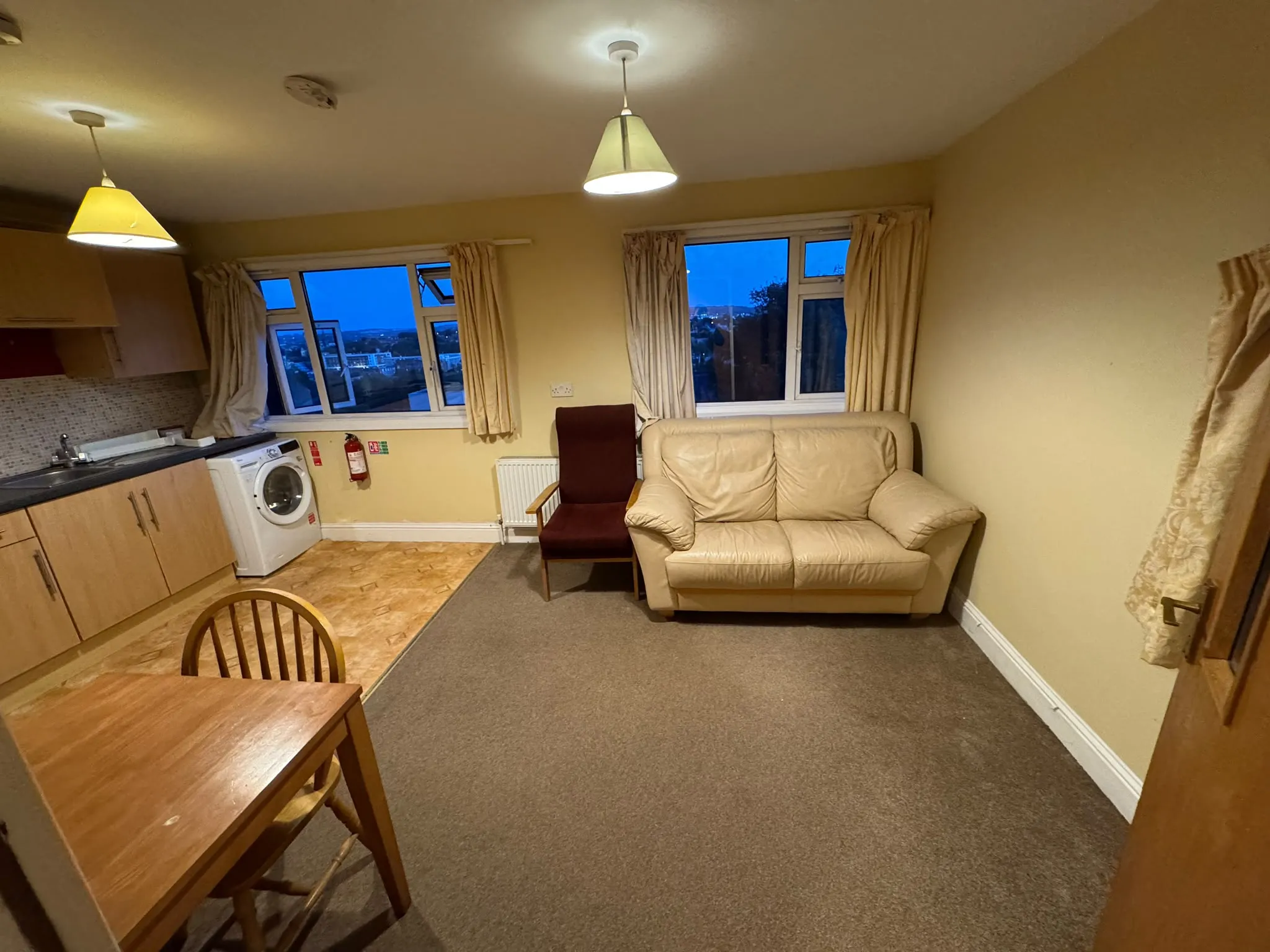 Open-plan living area with a small beige sofa and a single armchair, two windows with curtains, a dining table and chairs, kitchenette cabinets and a washing machine, and two ceiling pendant lights.