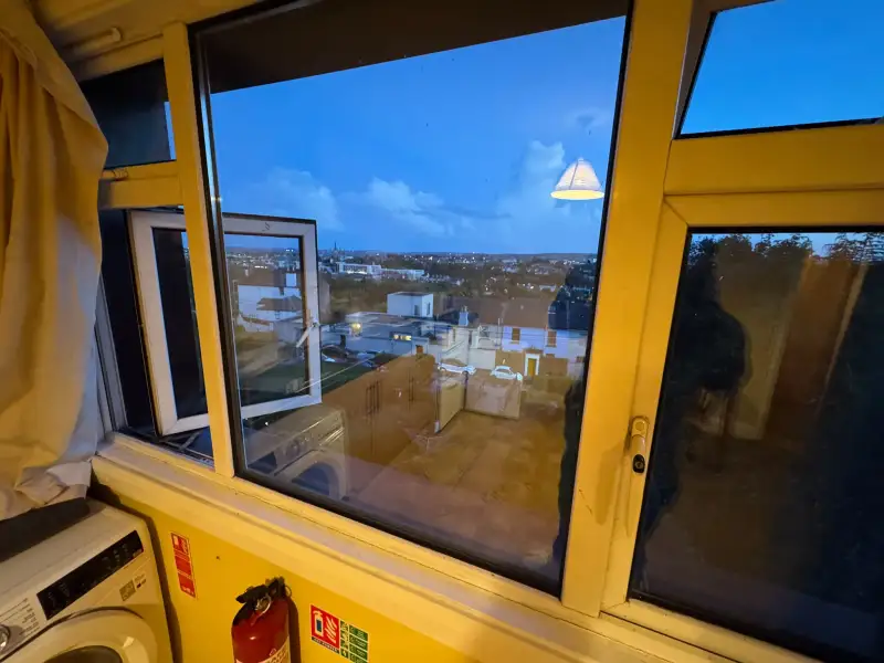 Interior view through a large window with an open casement showing a cityscape at dusk; curtain, washing machine and fire extinguisher visible.