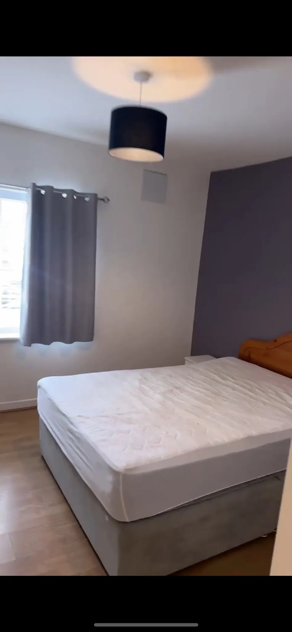Bedroom showing a double bed with mattress, wooden headboard, bedside table, window with grey curtain, and a ceiling pendant light.
