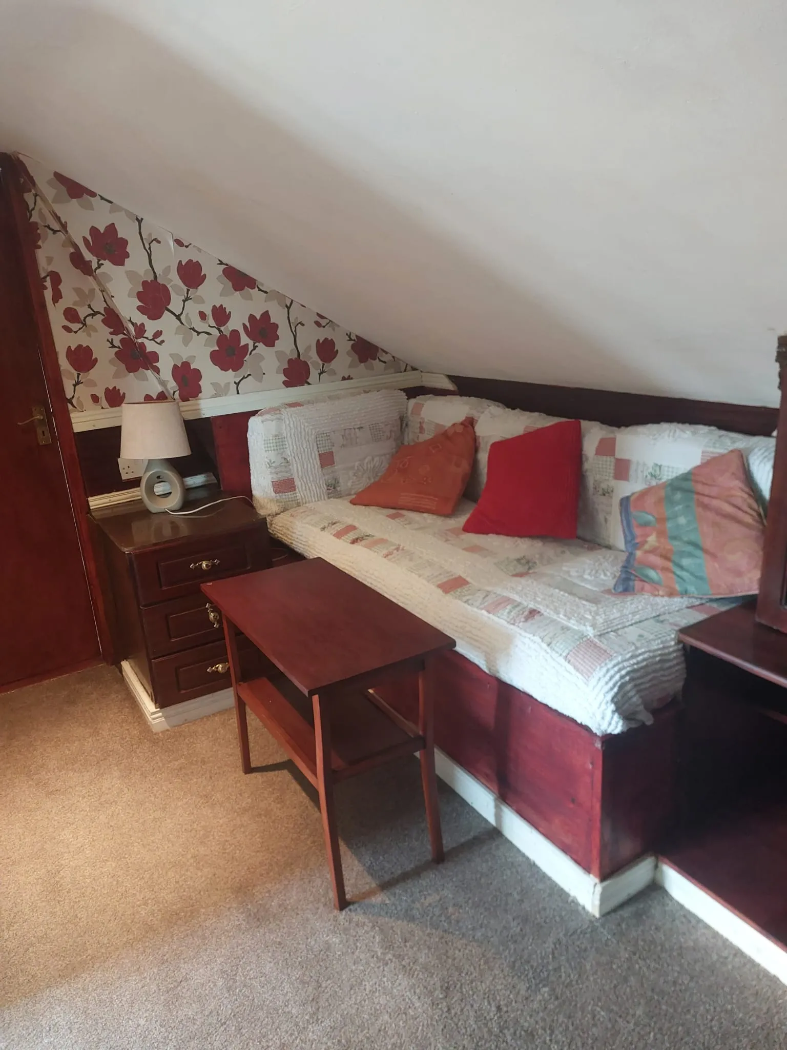 Bedroom with a low sloped ceiling, a daybed with patterned quilt and cushions, a bedside table with lamp, floral wallpaper on one wall, and a small wooden fold-out table on carpet.
