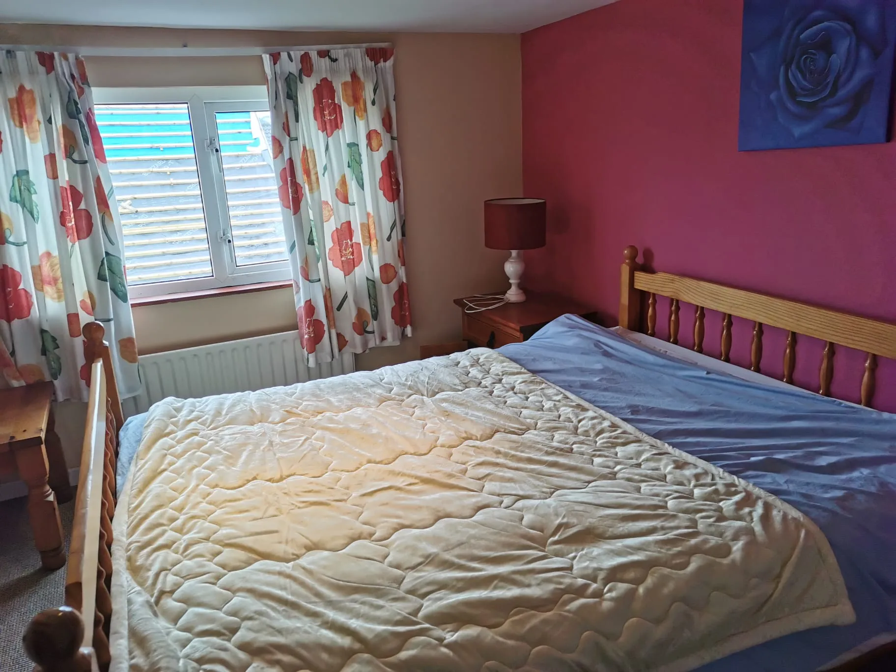 Bedroom with a double bed, wooden headboard, bedside table with a lamp, window with floral curtains and a radiator below.