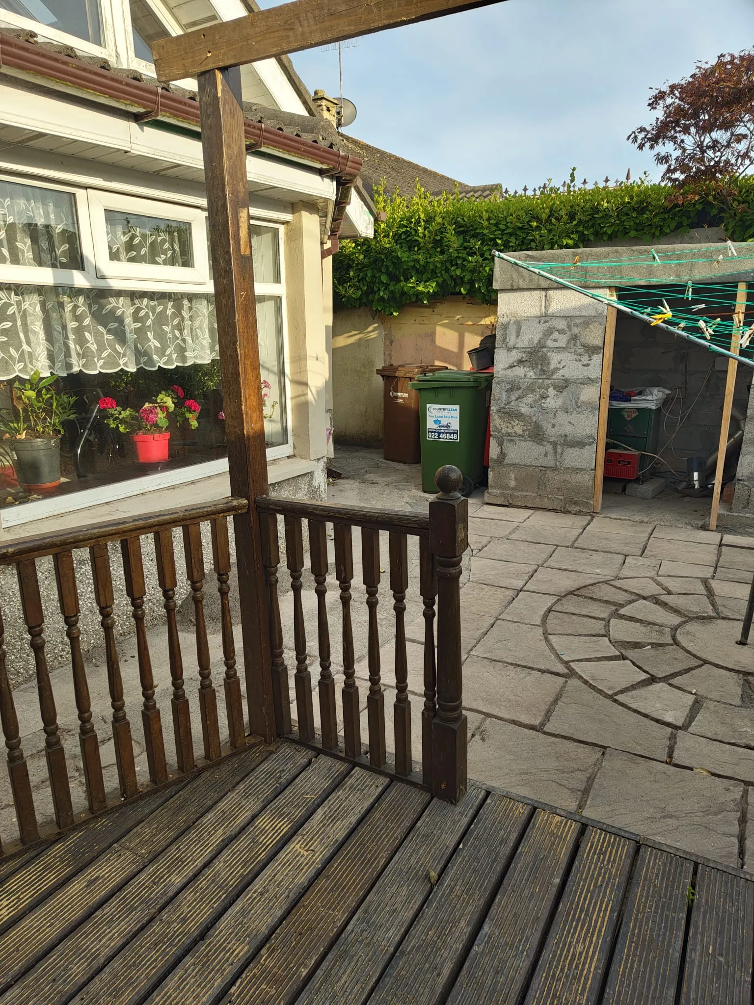 Outdoor patio with a wooden deck and railing, paved yard, a small concrete shed, clothesline, and two wheelie bins near a hedge.