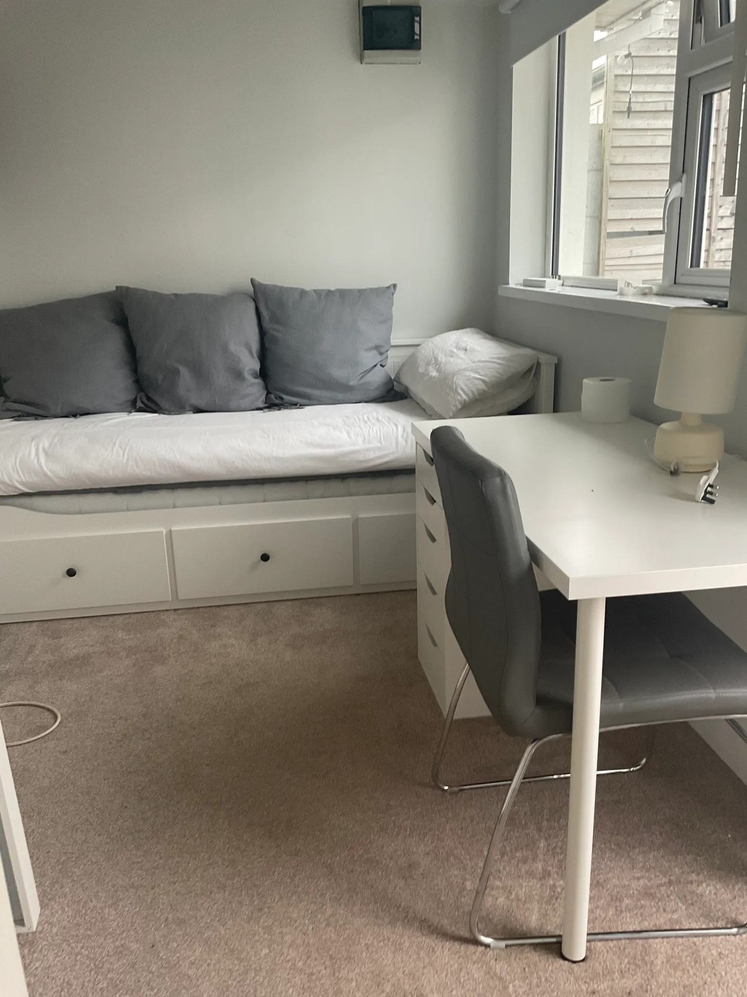Bedroom with a daybed against a wall with three gray cushions, a desk and chair by a window and a small table lamp.