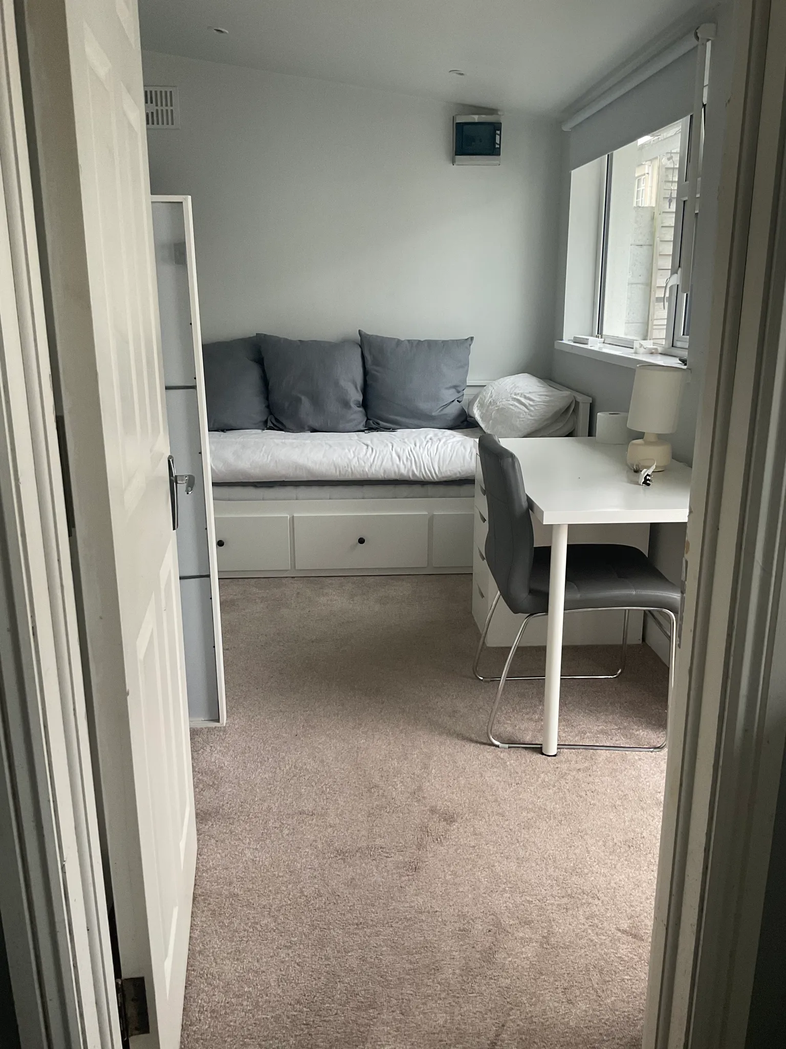 Bedroom with a built-in daybed with three gray cushions, a white desk and chair by a window with a roller blind, and drawers under the bed.