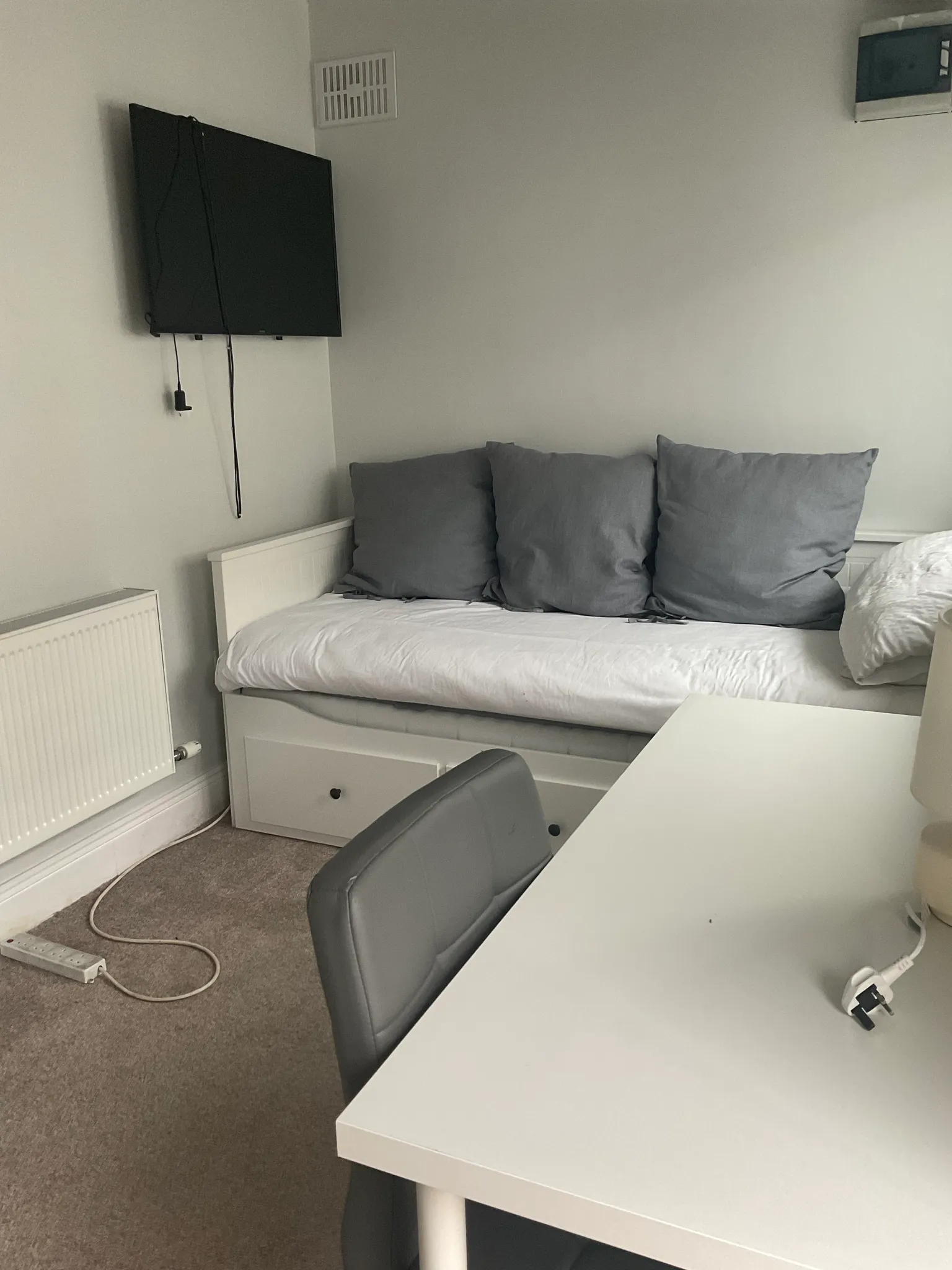 Bedroom with a single daybed with three grey pillows, a wall-mounted TV with visible cables, a white desk and grey chair, and a radiator.