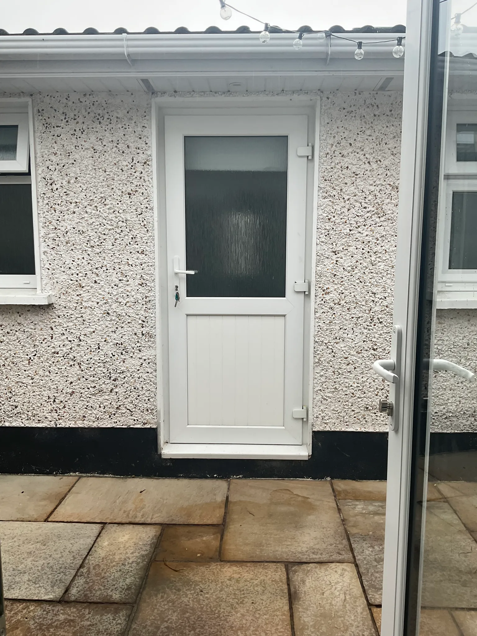 Exterior view of a white back door with frosted glass set in a pebble-dash wall, paved patio in foreground and a partially visible glass door frame at right.