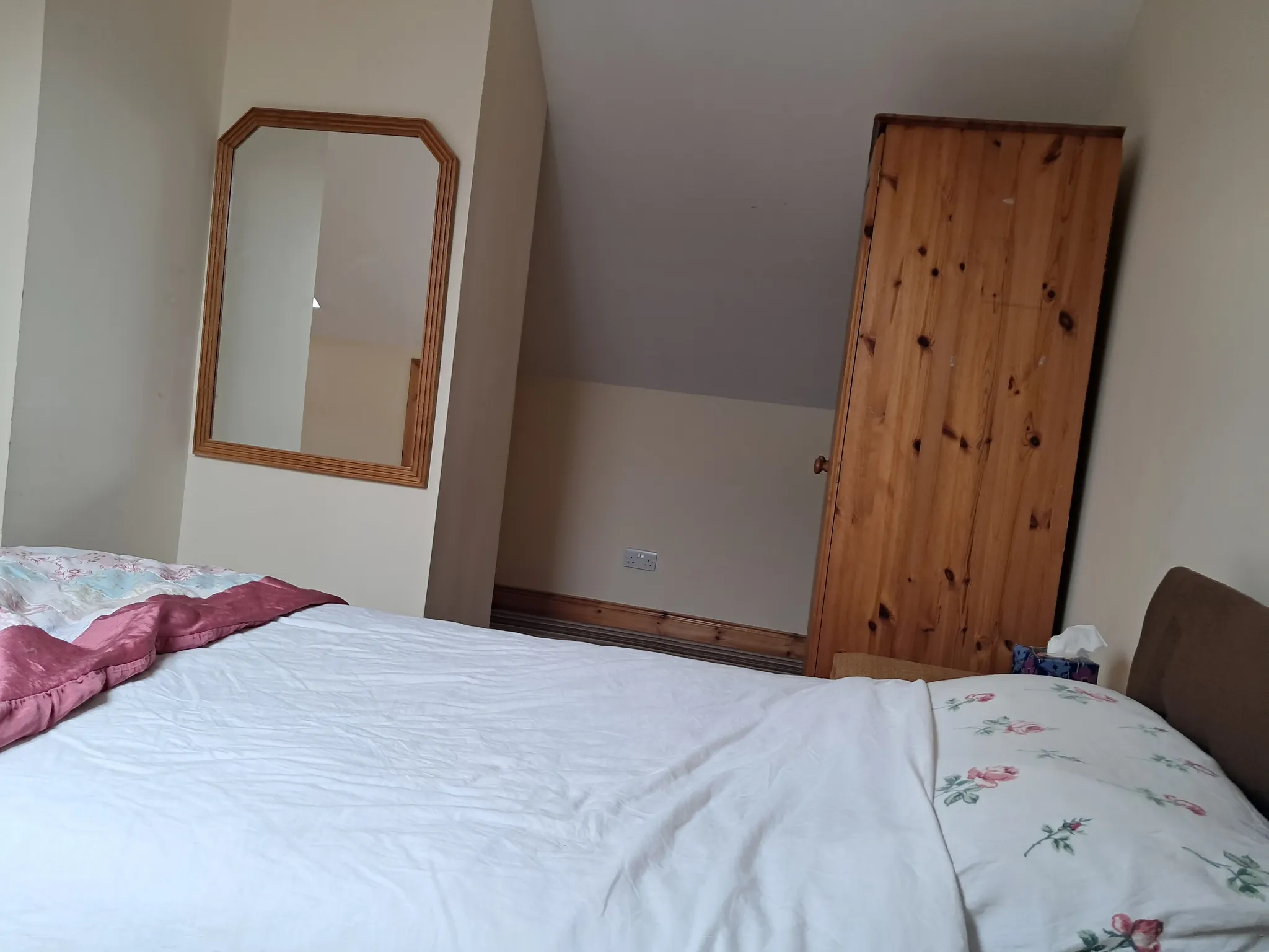 Bedroom showing a bed in the foreground, a wooden wardrobe on the right, a wall mirror and a small alcove with a power outlet.