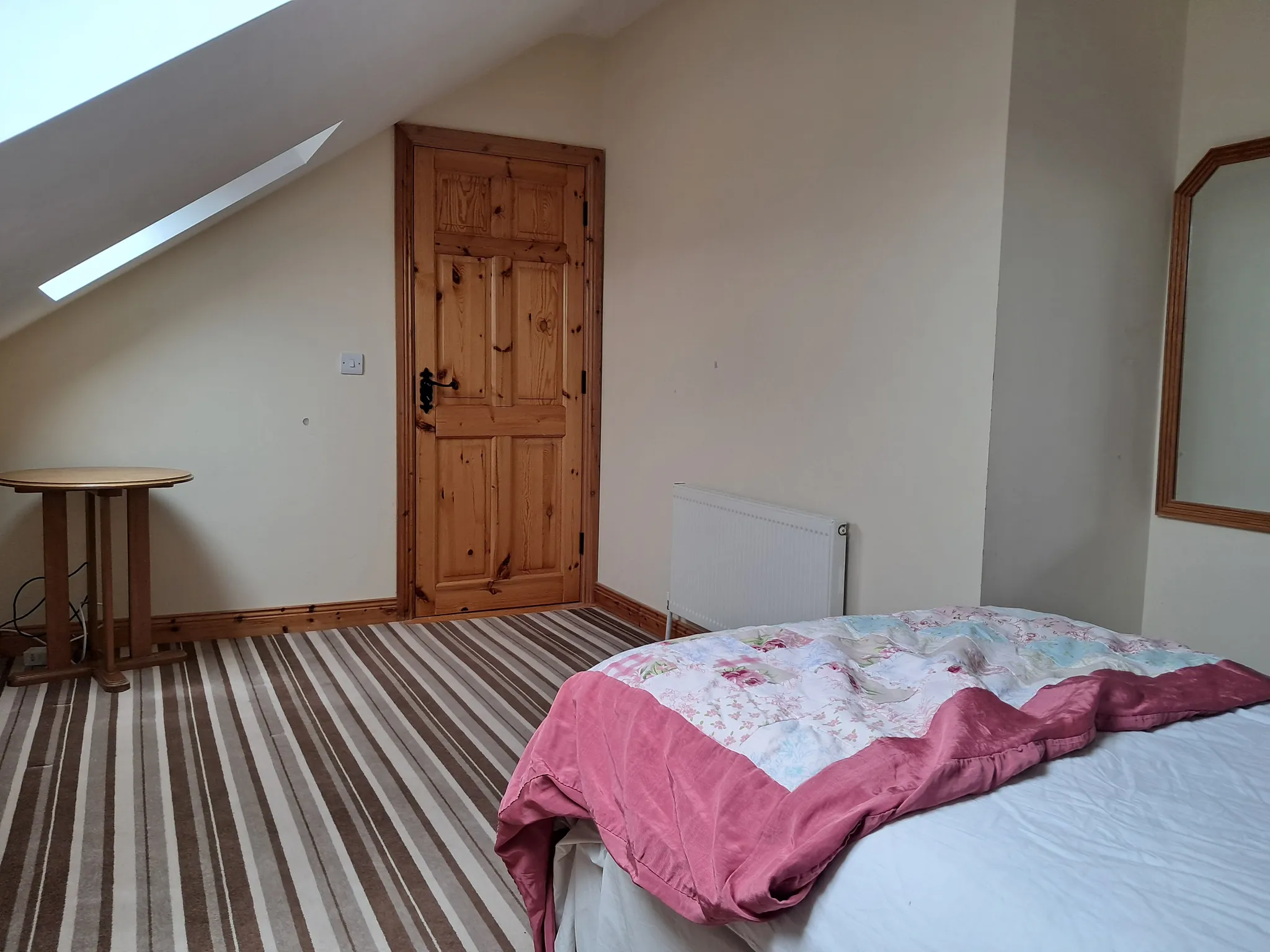 Bedroom with a wooden door, skylight, radiator, striped carpet and part of a bed with a pink quilt.