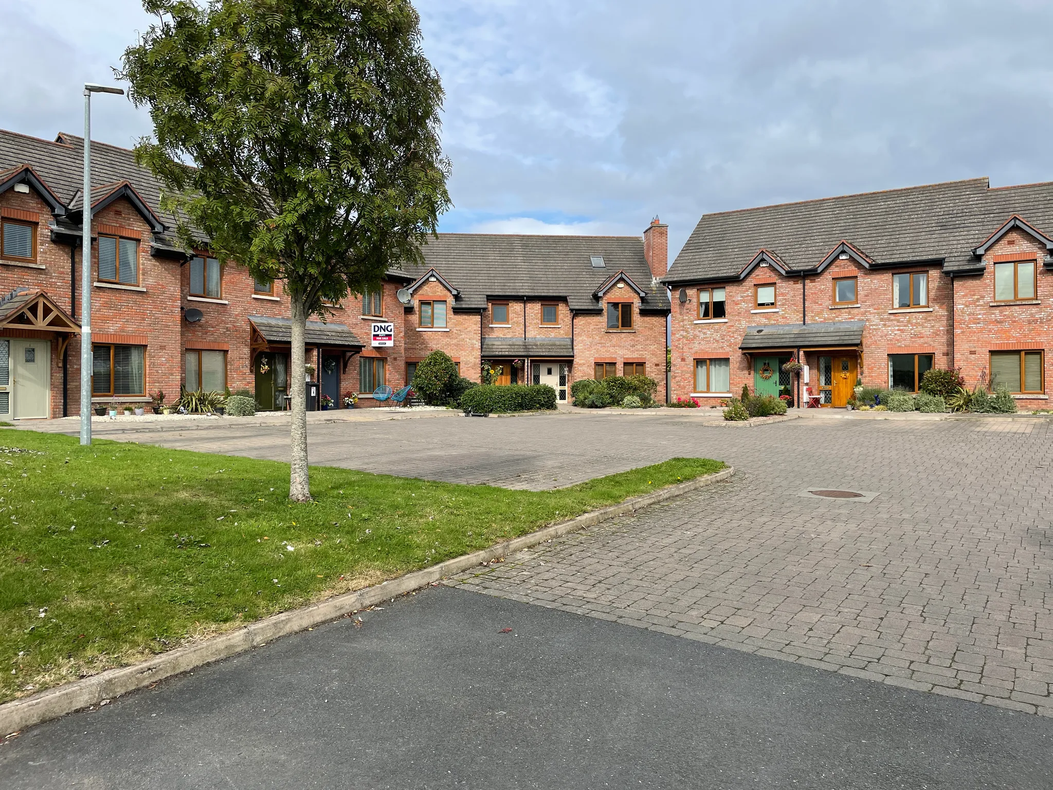 Exterior view of a row of brick townhouses with pitched roofs, arranged around a paved courtyard with green lawn and mature trees