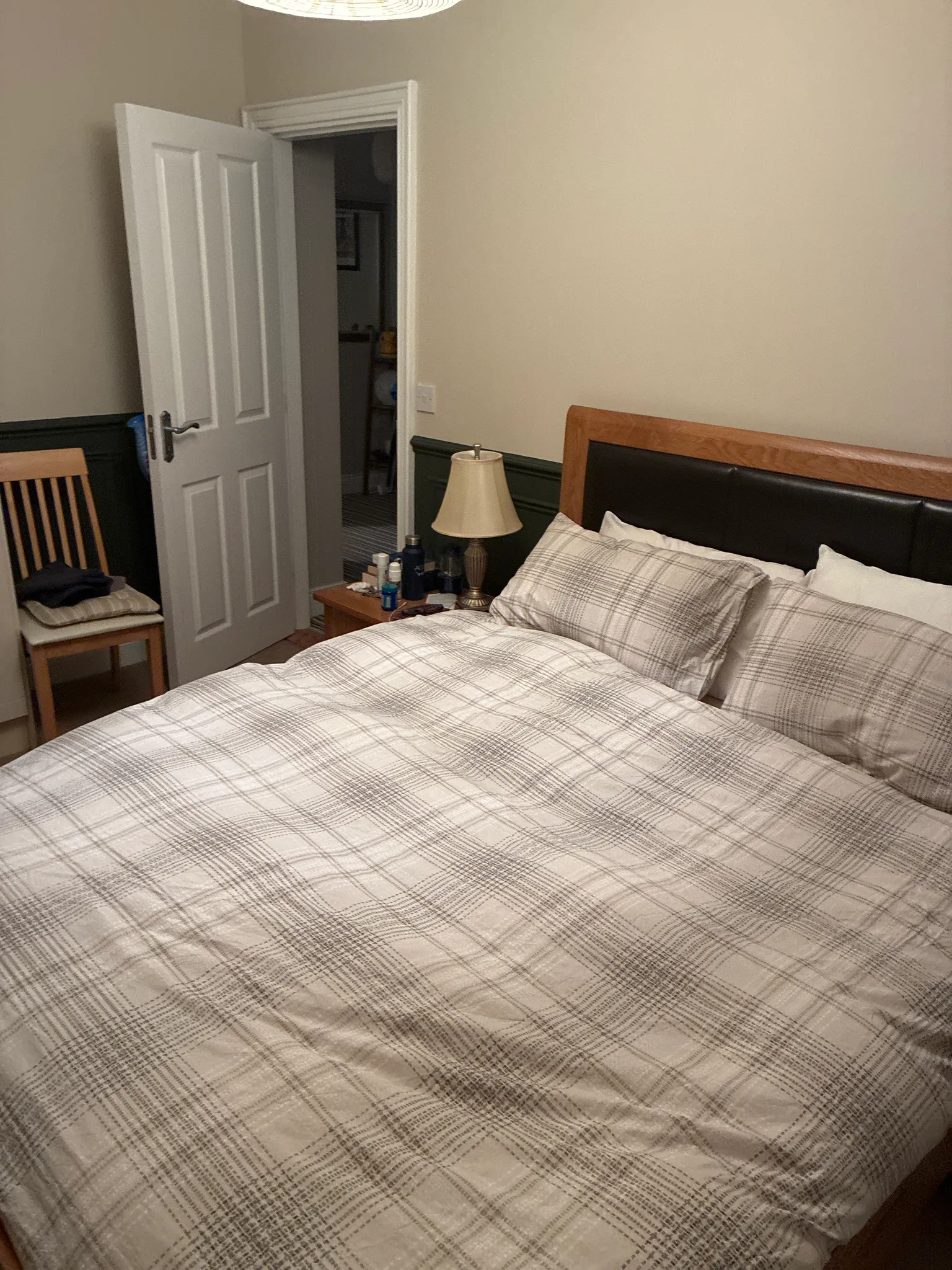 Bedroom with a double bed with plaid bedding, wooden headboard with black upholstery, bedside table with lamp, wooden chair, white closet door, and beige walls with dark wainscoting.