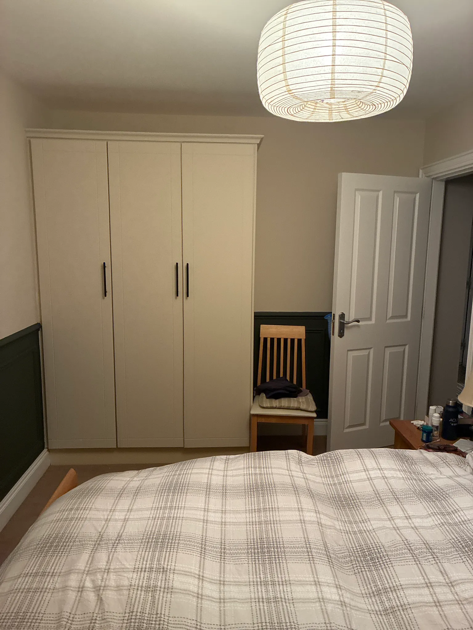Bedroom with double bed, large cream wardrobe with black handles, pendant ceiling light, white door, and wooden chair with cushion