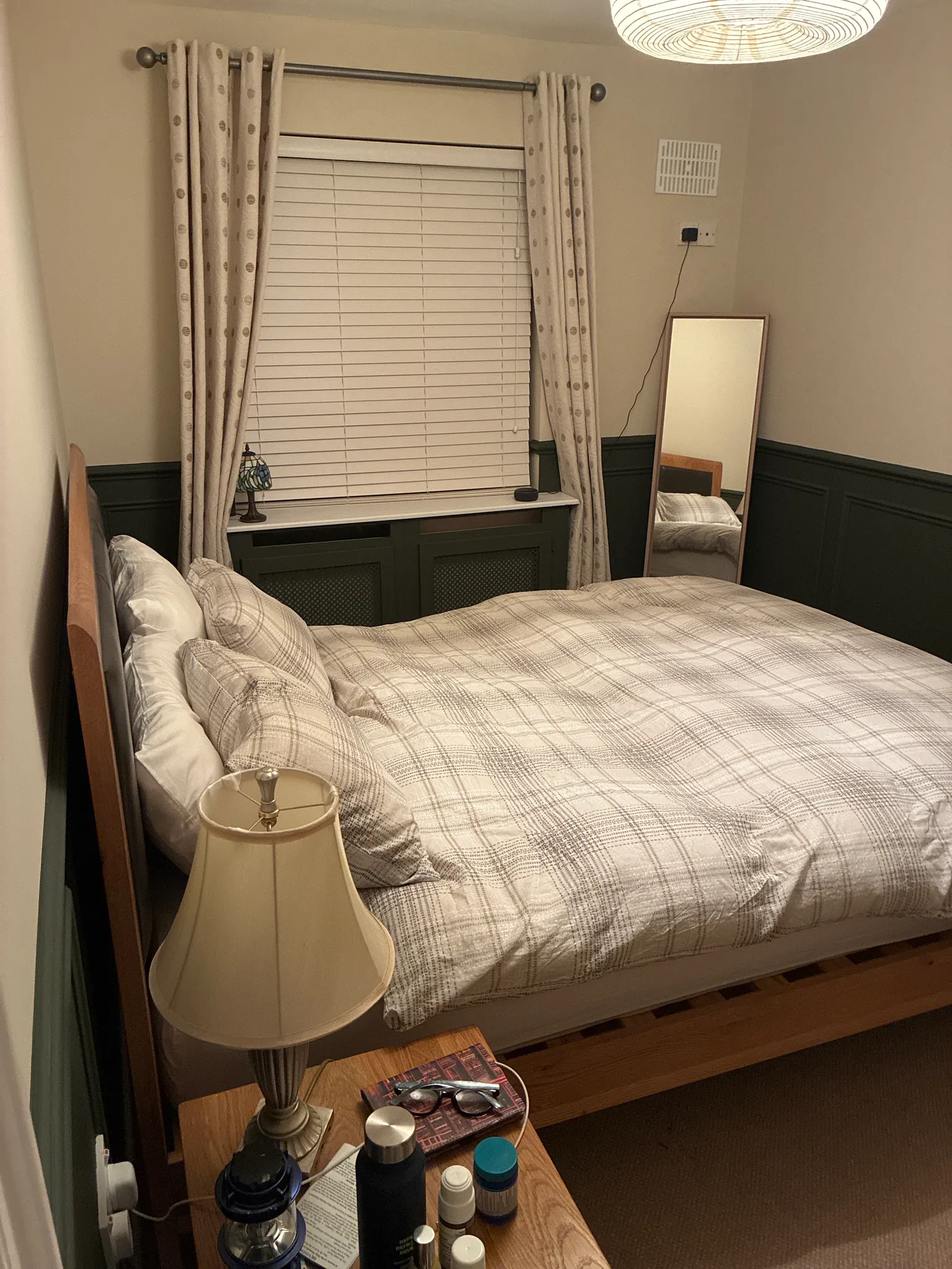 Bedroom with a double bed, plaid bedding, and pillows. Window with horizontal blinds and cream curtains. Black wainscoting on walls, ceiling fan, bedside lamp, and wooden nightstand with personal items.