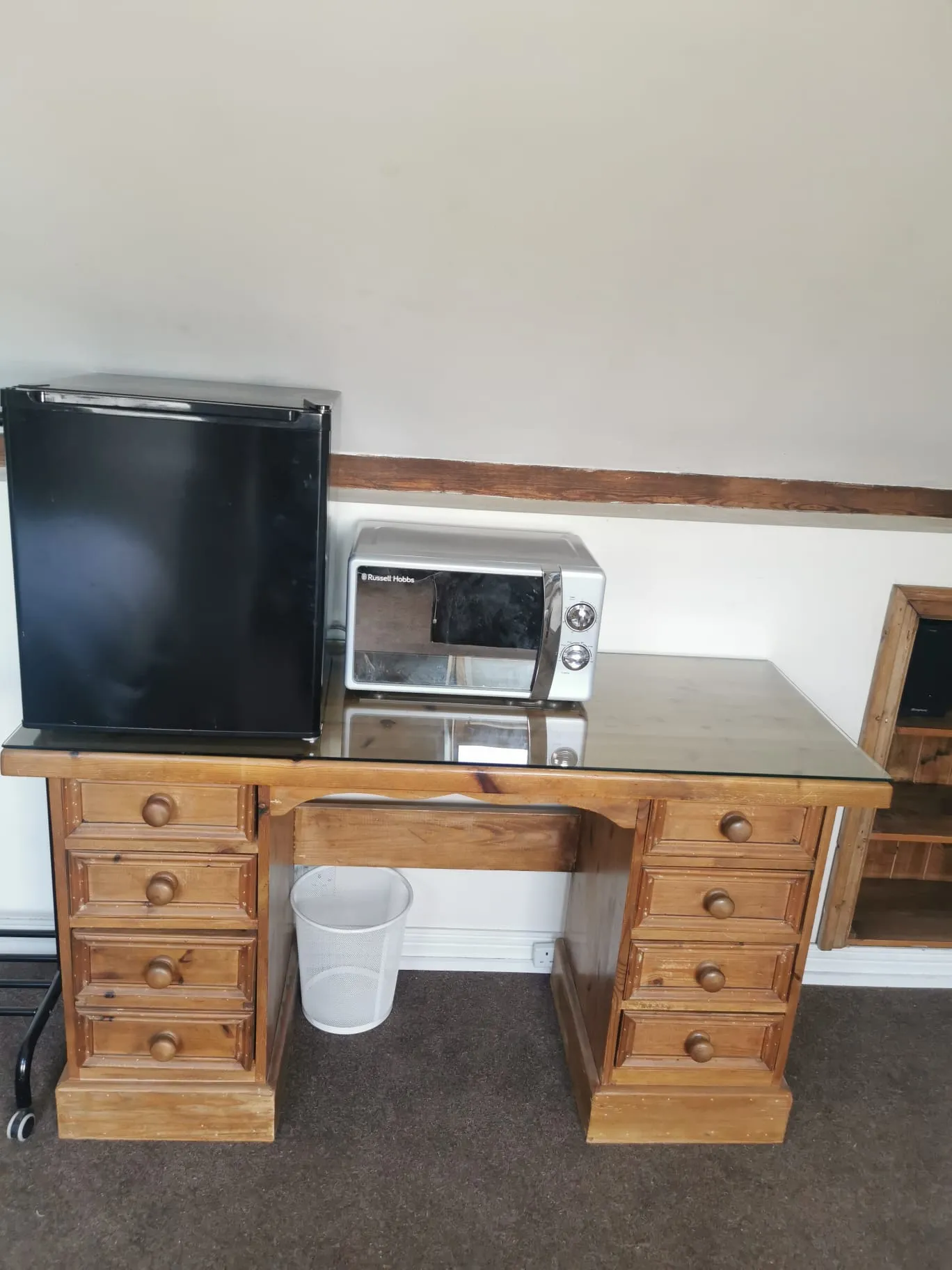 Wooden desk with six drawers, a small black mini fridge and a silver microwave on top, and a white waste bin underneath against a plain wall.