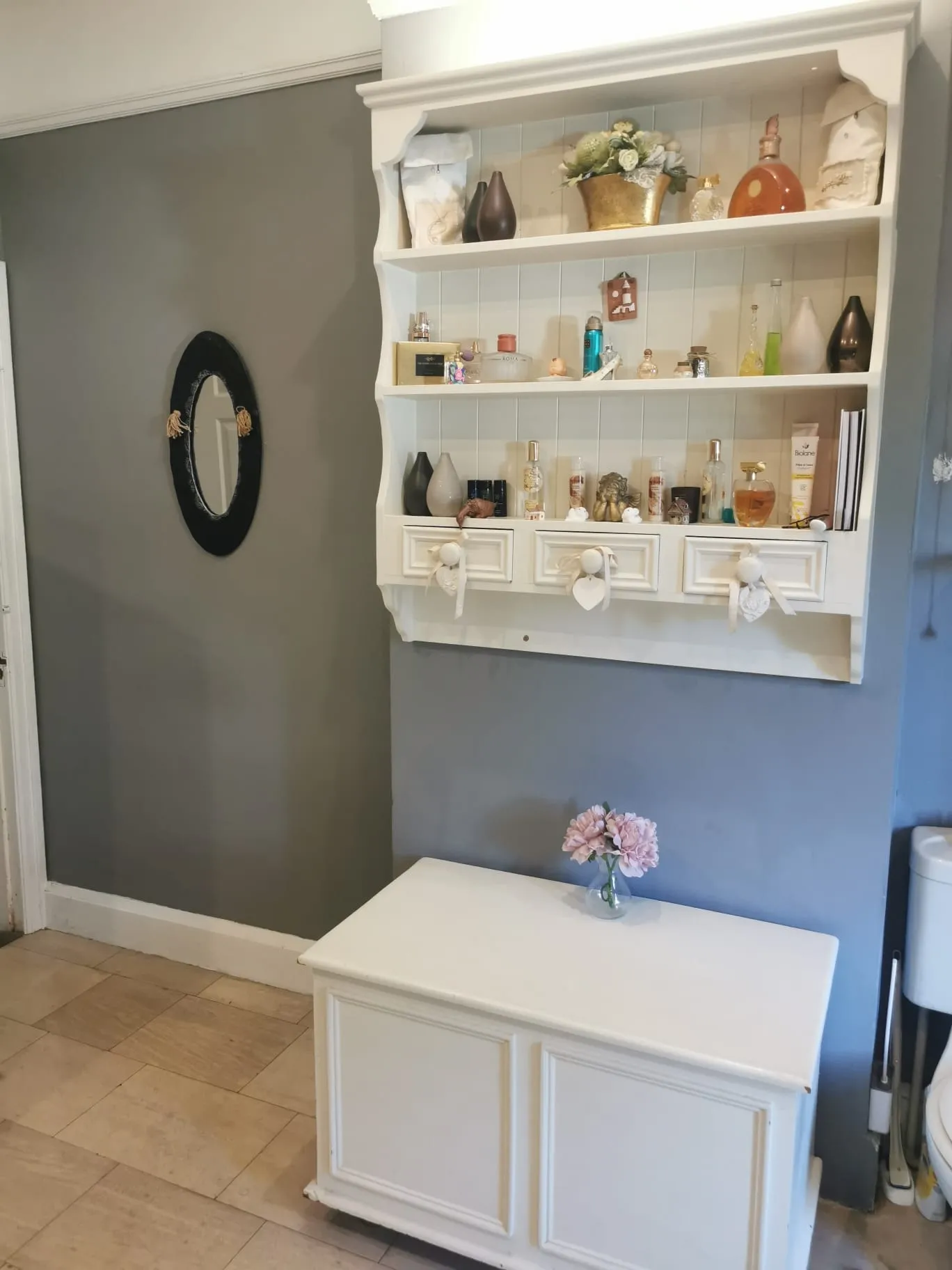 Bathroom with a wall-mounted white shelving unit holding toiletries and decorative items, a small white cabinet with a vase of pink flowers, and an oval wall mirror on a grey wall.