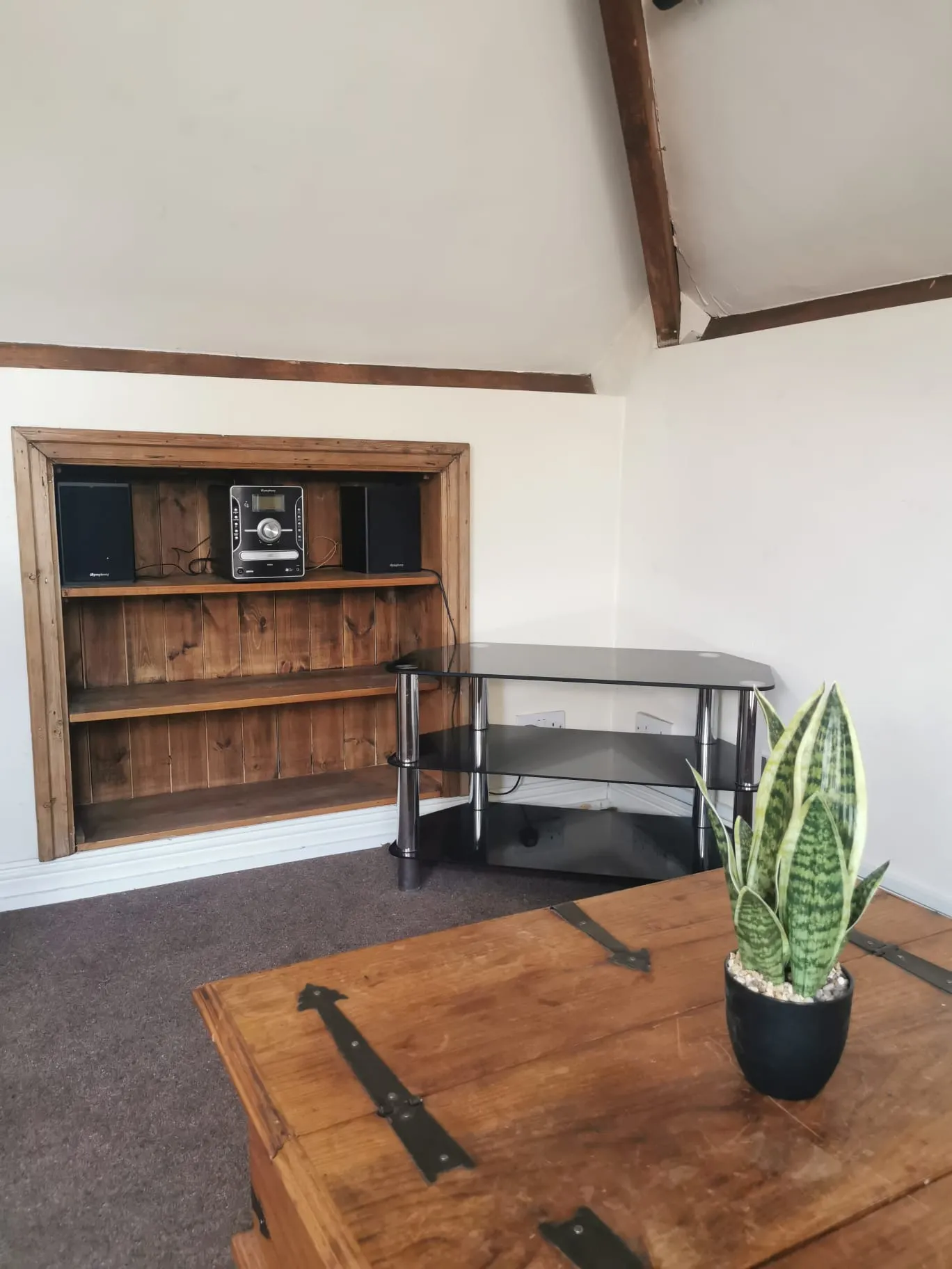 Living room corner with a built-in wooden shelving niche holding a stereo and speakers, a glass corner TV stand, a wooden chest coffee table and a potted snake plant; ceiling beam visible.