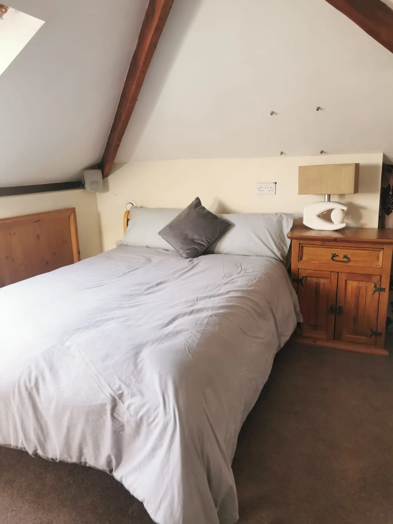 Bedroom with a double bed, gray bedding and a single decorative pillow, wooden bedside cabinet with a lamp, sloped ceiling with exposed wooden beams