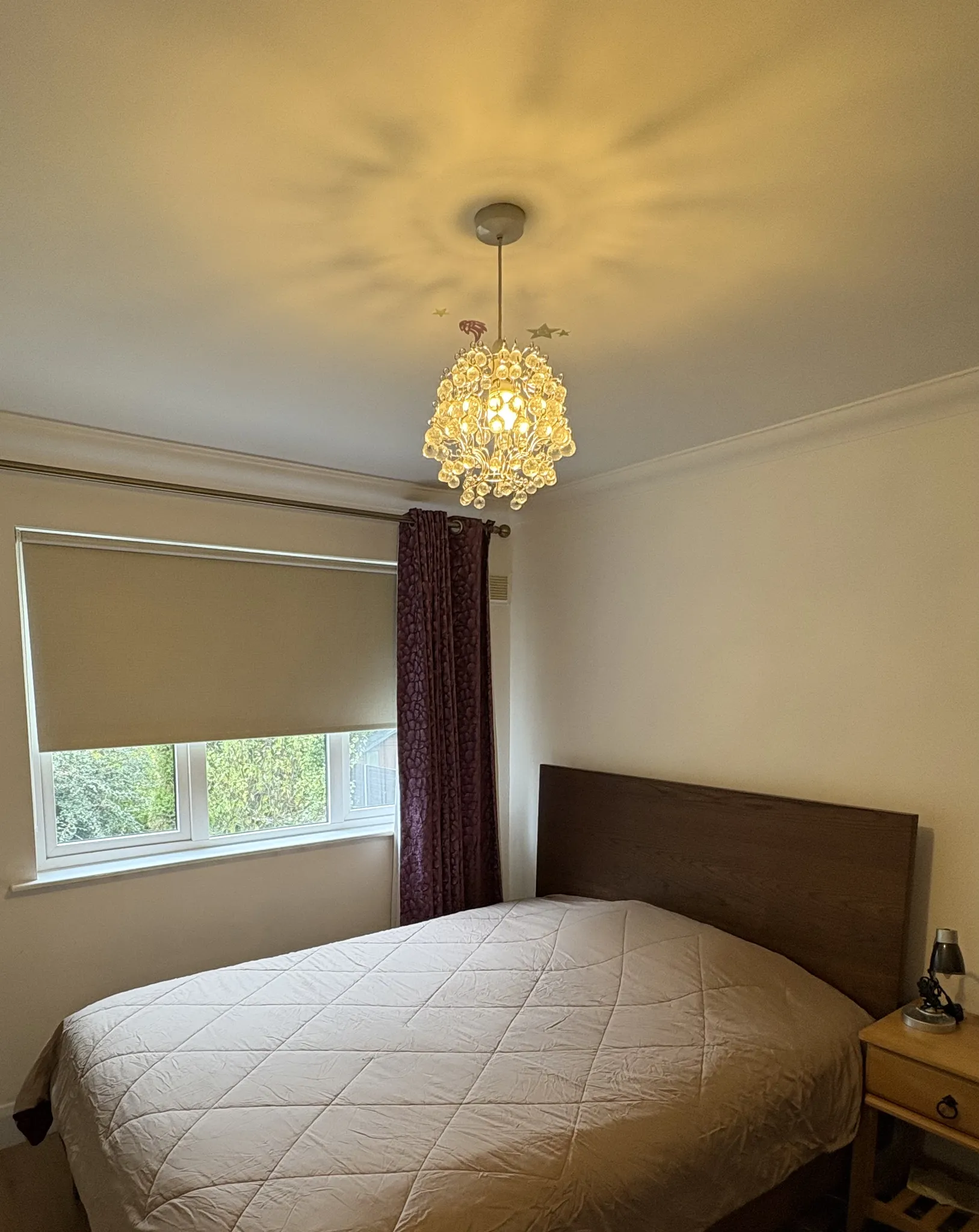 Bedroom with a double bed, wooden headboard, window with a roller blind and purple curtain, pendant chandelier, and a bedside table with a lamp.