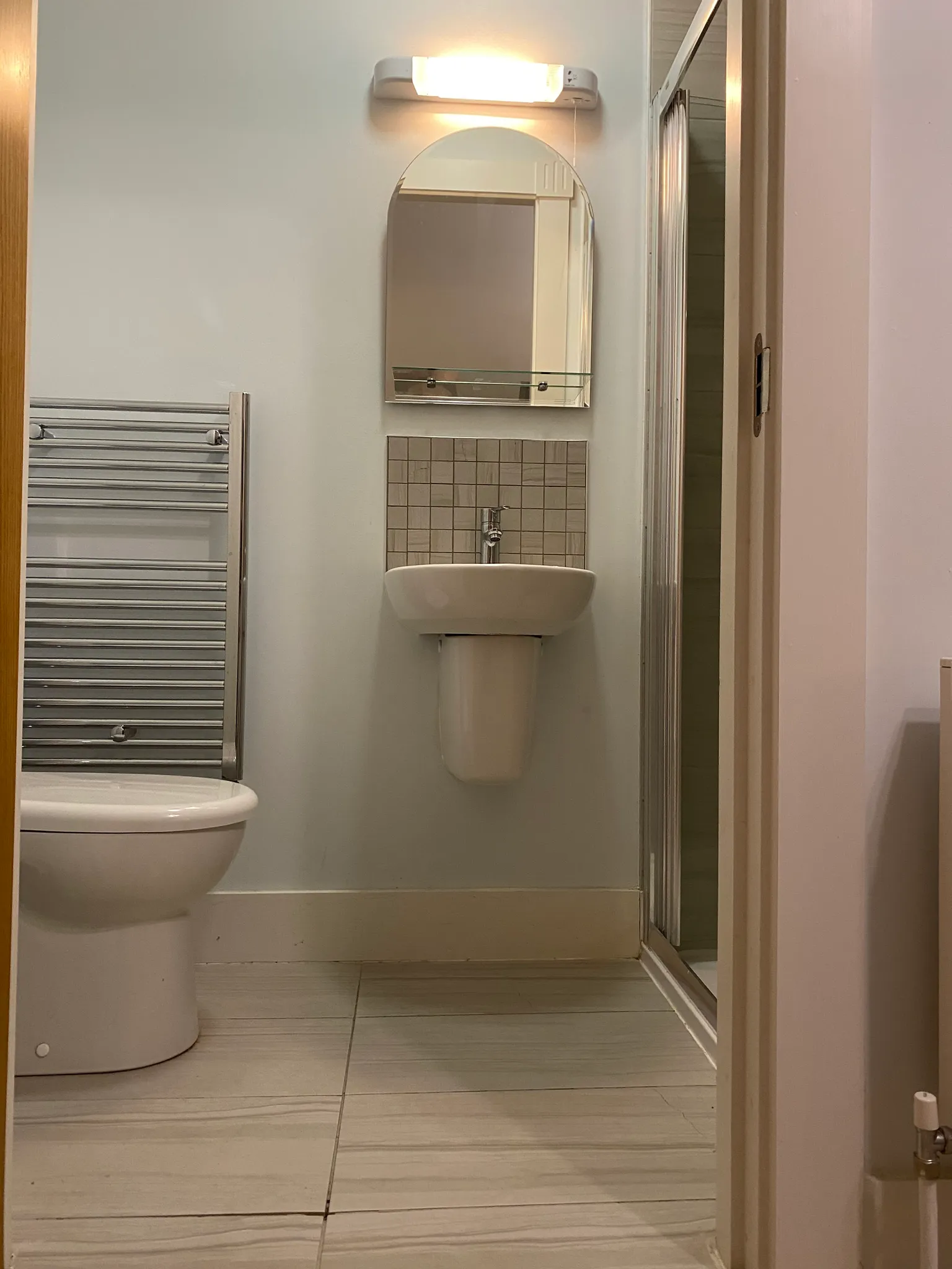 Bathroom with a wall-mounted sink, arched mirror and light above, towel radiator, toilet bowl at left and shower doorway at right.