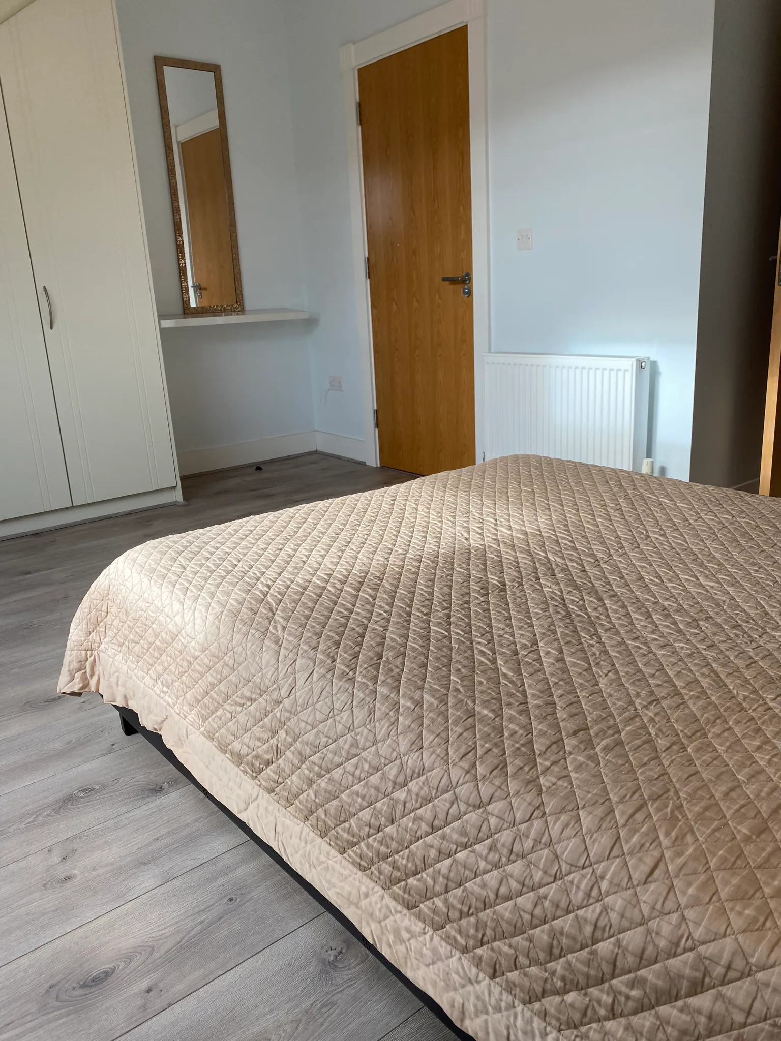 Bedroom with a quilted bedspread in foreground, wardrobe, wall mirror, wooden door and a radiator