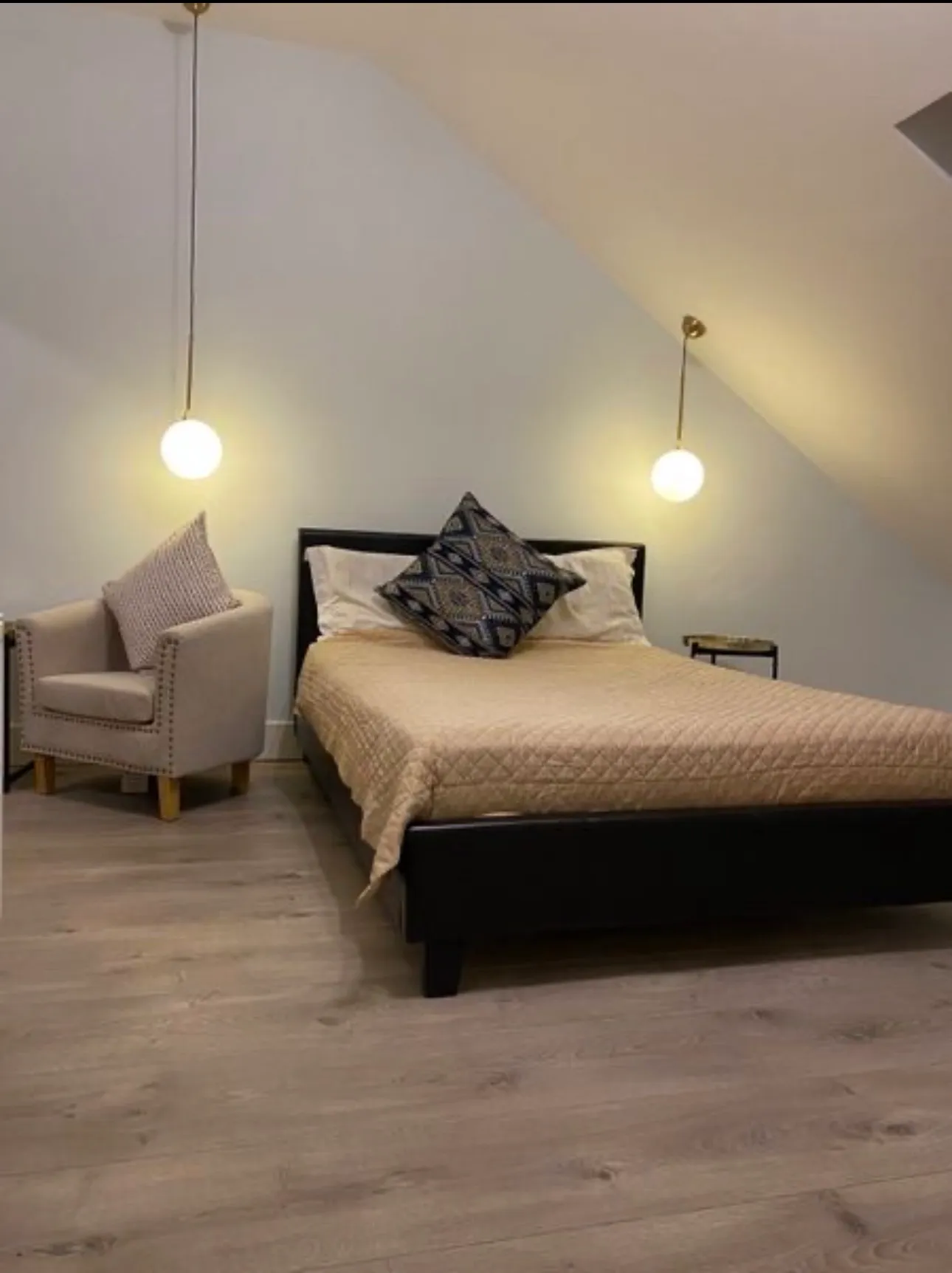 Bedroom with a double bed, patterned throw pillow, two hanging pendant lights, an armchair with a cushion, and a small side table; sloped ceiling and wood-look floor visible.