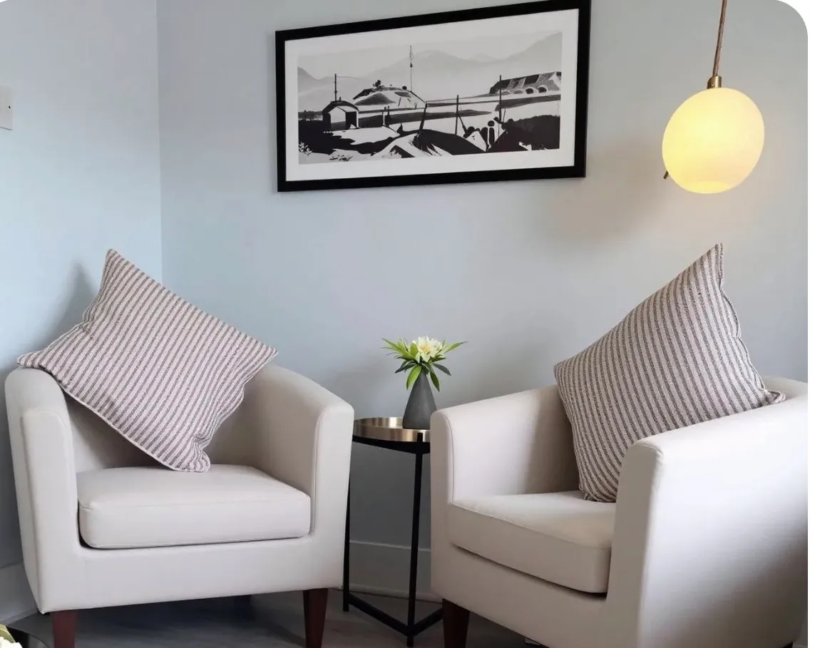 Two upholstered armchairs with striped throw pillows facing a small round side table holding a vase of flowers, framed wall art above, and a hanging round lamp to the right.