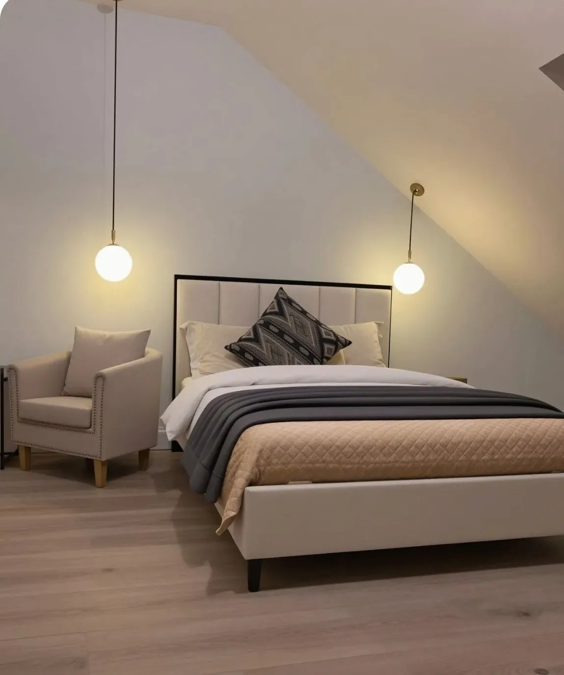 Bedroom with a double bed, patterned throw pillow, armchair to the left, two hanging pendant lights, and a sloped ceiling.