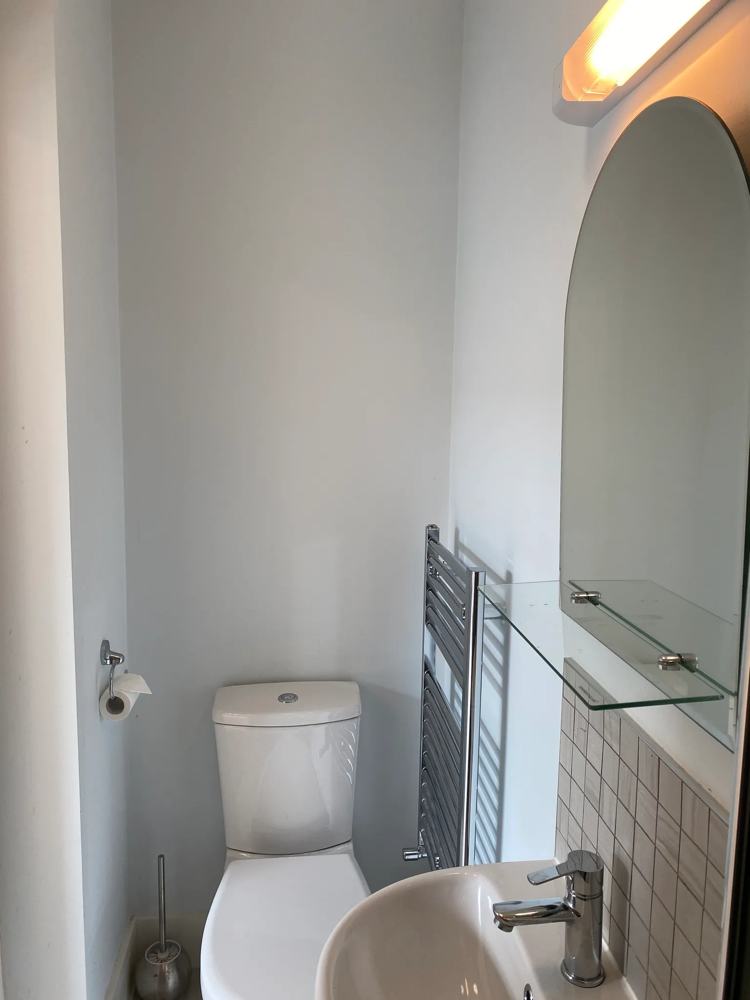 Small bathroom showing a toilet, sink with faucet, wall-mounted glass shelf and arched mirror, vertical heated towel rail, and toilet paper holder.