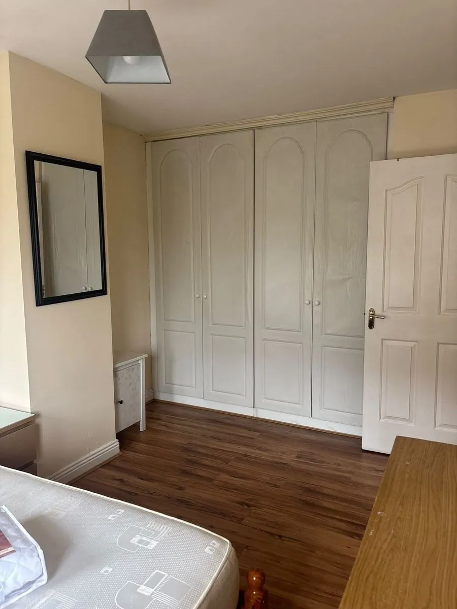 Bedroom with built-in white wardrobes, wooden floor, a mattress in foreground, wall mirror, bedside table and a partially open door.