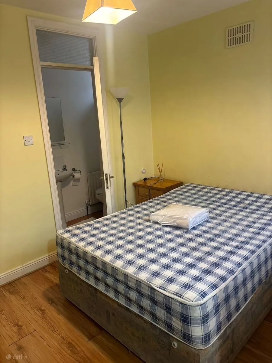 Bedroom with a double mattress on a divan base, checkered mattress cover, small wooden nightstand, tall floor lamp, and an open door leading to a small bathroom