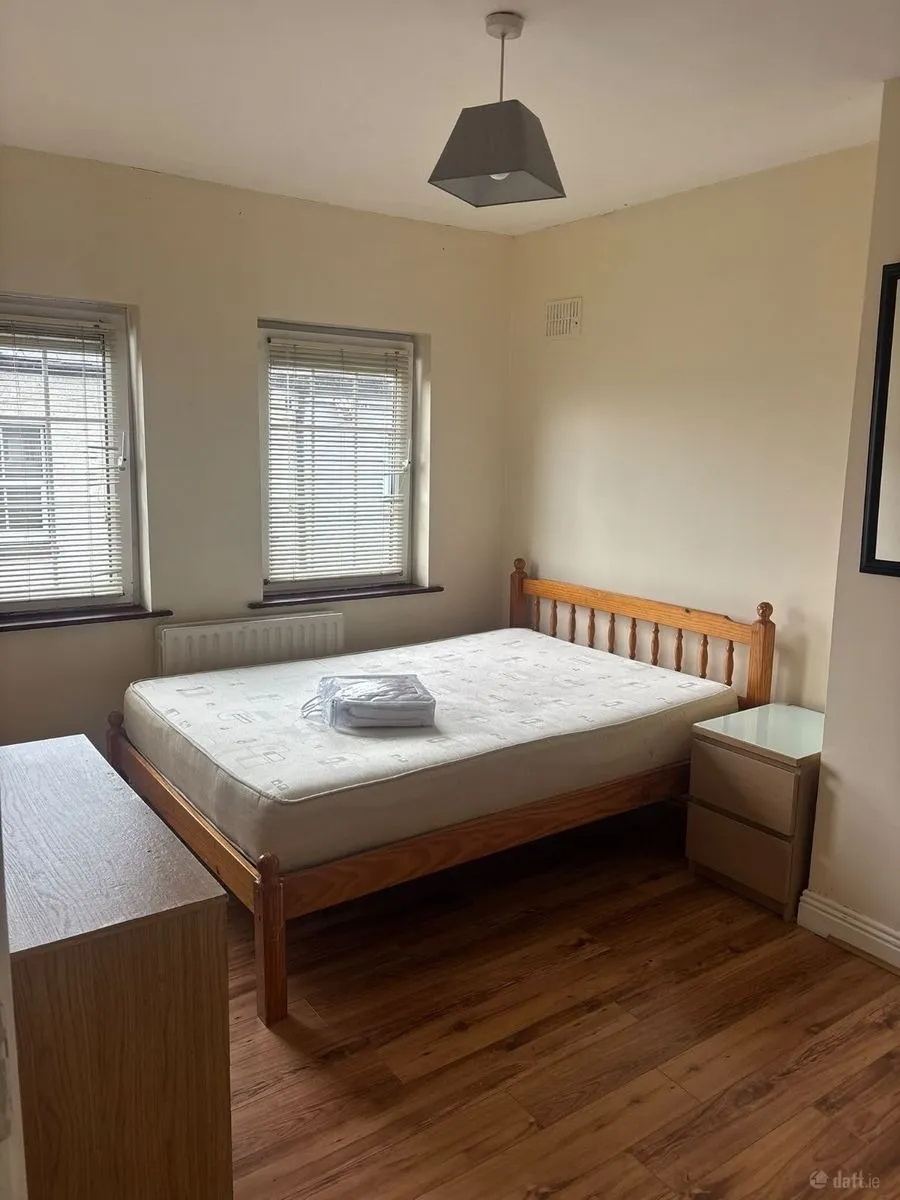Bedroom with a wooden double bed and mattress, folded linens on the mattress, two windows with blinds, bedside table and chest of drawers, pendant ceiling light and wooden floor.