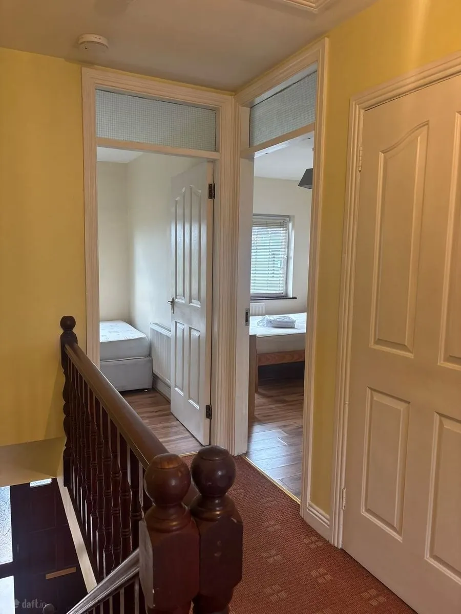Hallway landing with carpeted floor and wooden banister, open doors leading to two small bedrooms with visible beds and a window
