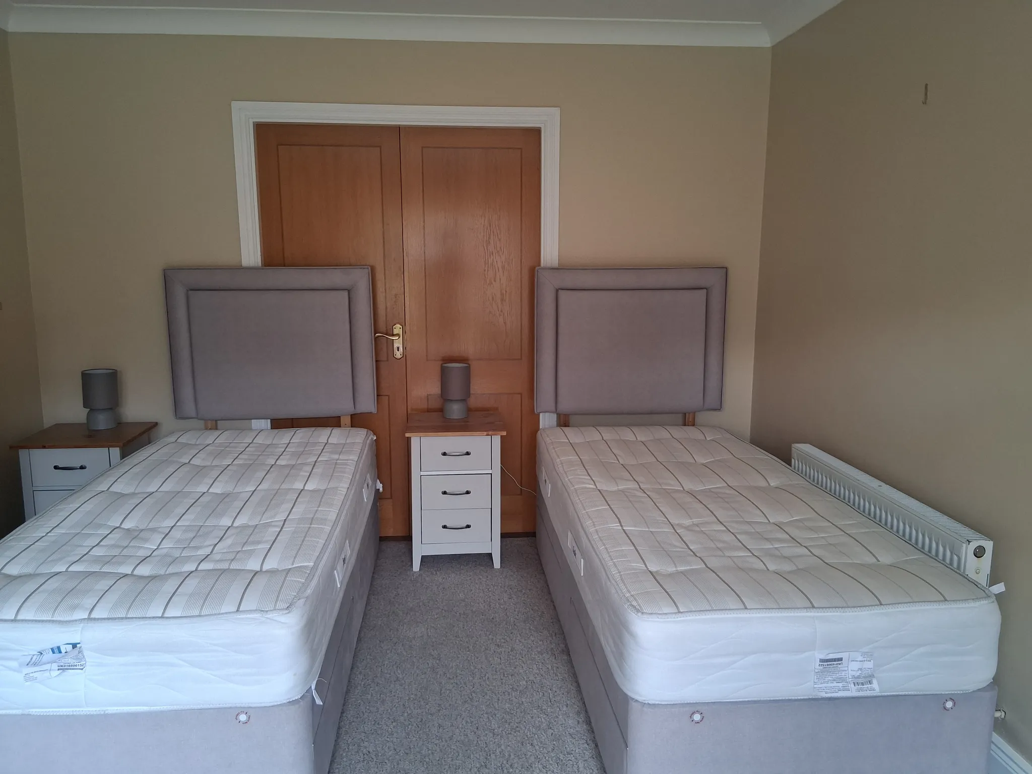 Bedroom with two single beds separated by a small white bedside drawer, padded headboards, a central wooden door and two small bedside lamps.