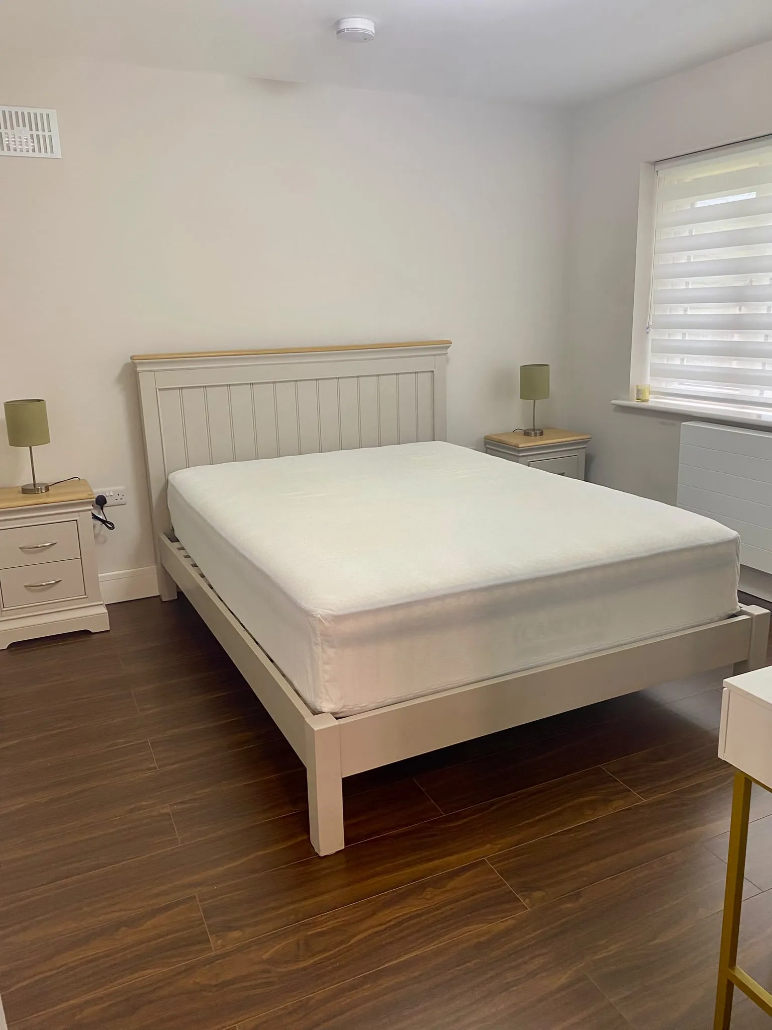 Bedroom with a bed and mattress, two bedside tables with lamps, a window with roller blinds, and wood-look flooring.