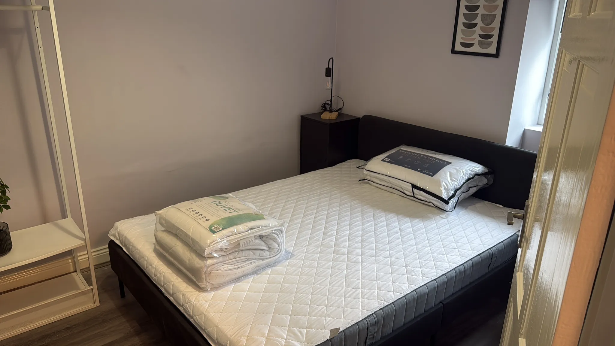 Bedroom with a double bed and mattress, packaged pillows and bedding on the bed, a small nightstand with a lamp, a window and framed art on the wall.