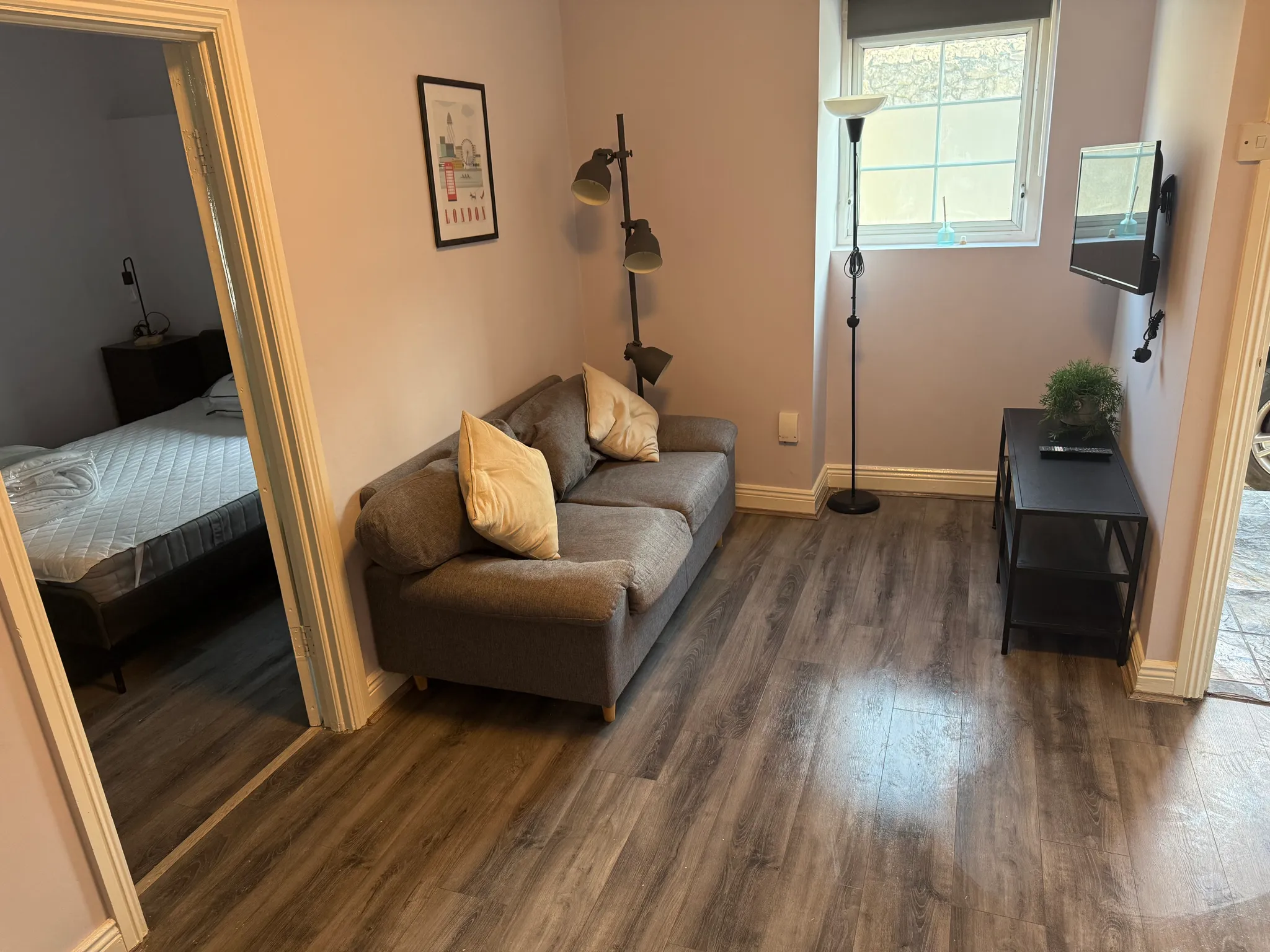 Small living room with a grey two-seater sofa with cushions, tall floor lamps, a window, wall-mounted TV and a small black side table; doorway at left shows a bedroom with a mattress.