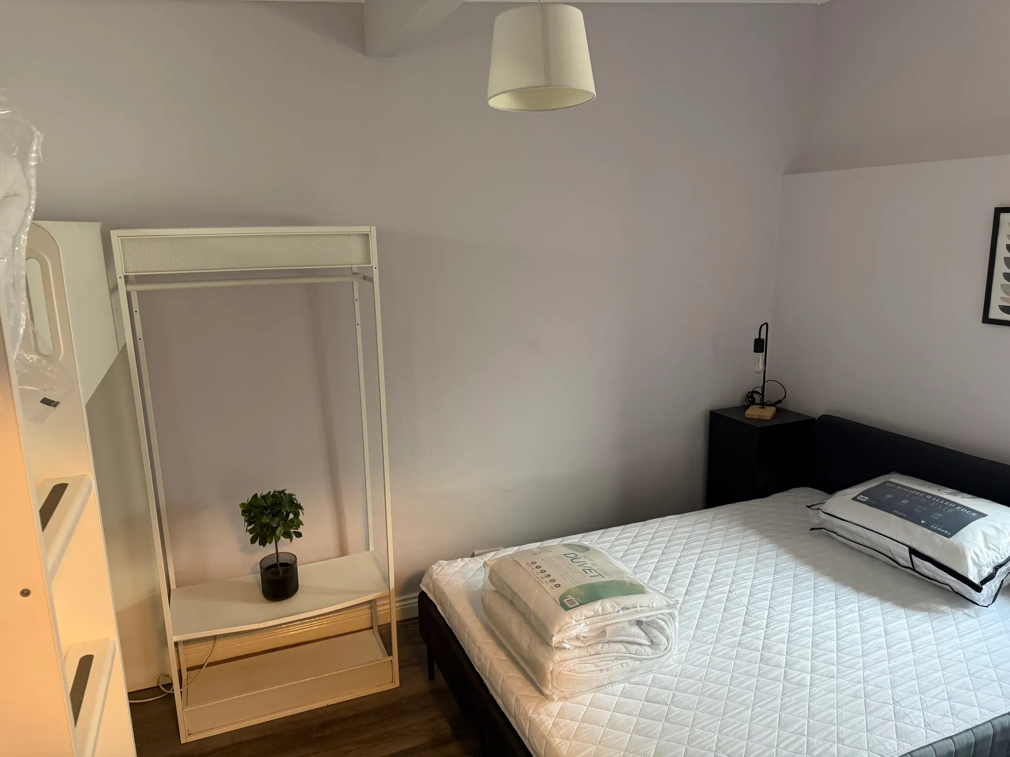 Bedroom showing a mattress on a bed frame with folded bedding on top, an open metal clothing rack and shelf with a small potted plant, a bedside table with a lamp, and a ceiling light.