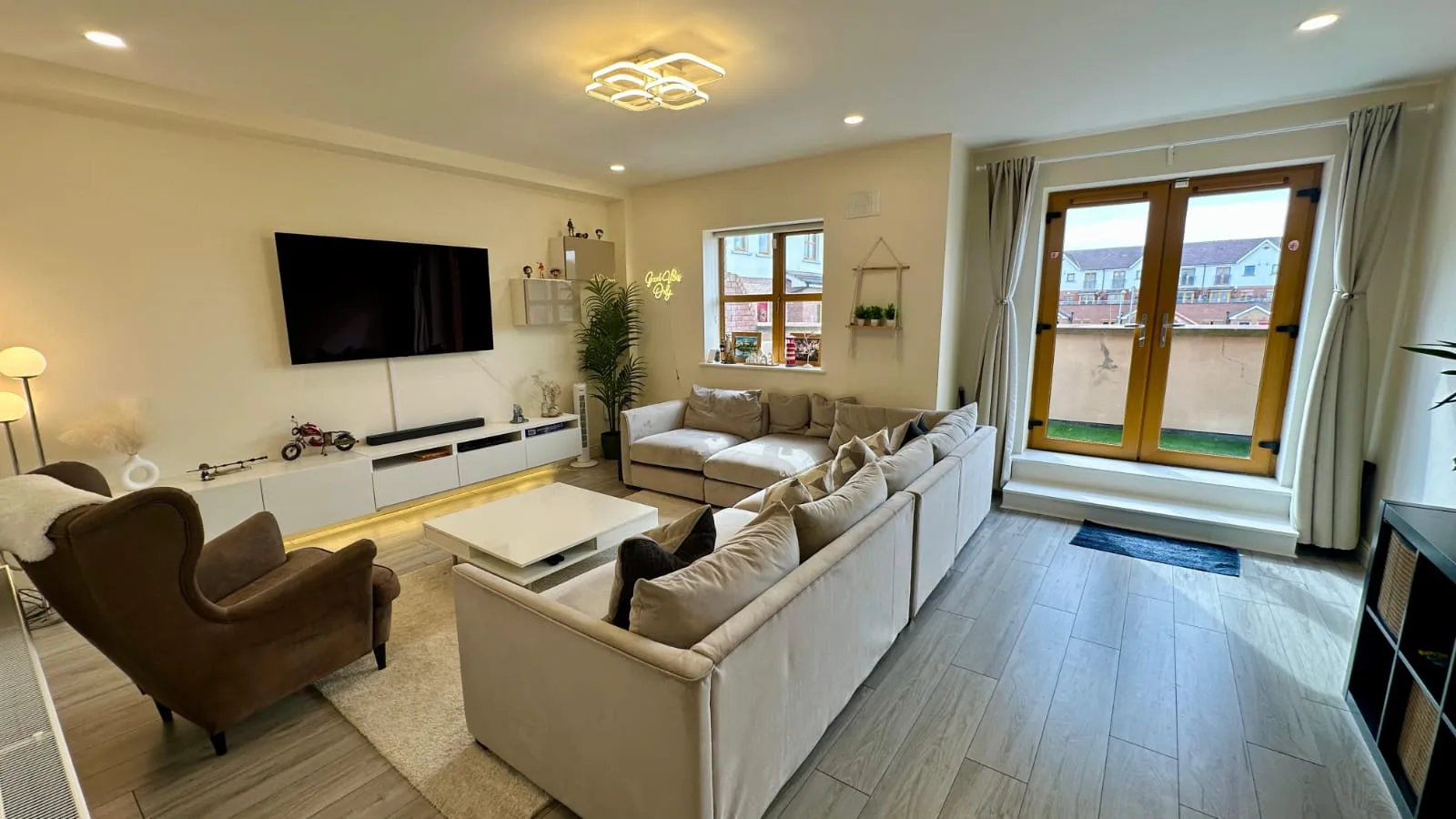 Living room with a large L-shaped sofa, coffee table, wall-mounted TV, armchair, and glass double doors to a small balcony or exterior