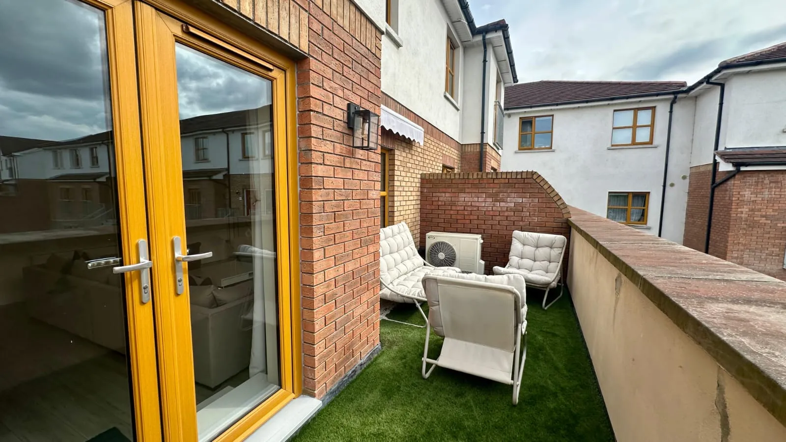 Balcony with four cushioned outdoor chairs on artificial grass, brick walls, an air conditioning unit, and yellow-framed glass patio doors.
