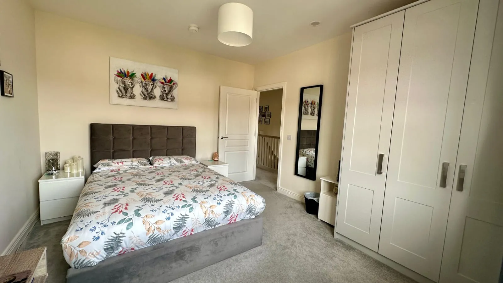 Bedroom with a double bed with patterned duvet, upholstered headboard, two bedside tables, tall white wardrobe, full-length mirror and ceiling light.