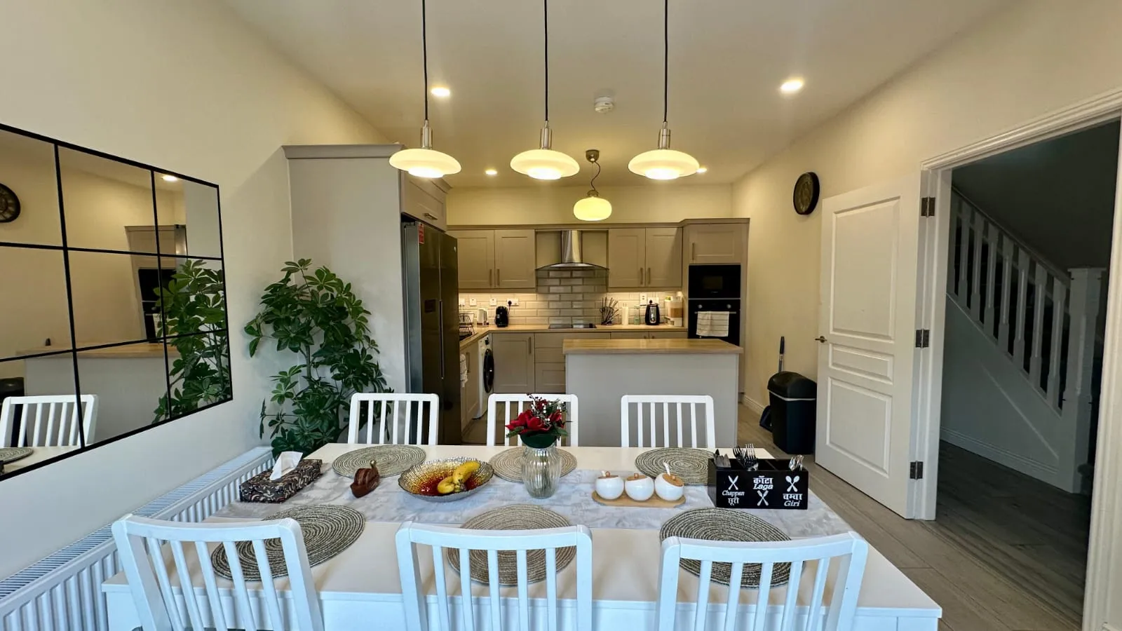 Dining room with a rectangular table set for six, a potted plant, wall mirror, and an open-plan kitchen with island and pendant lights.