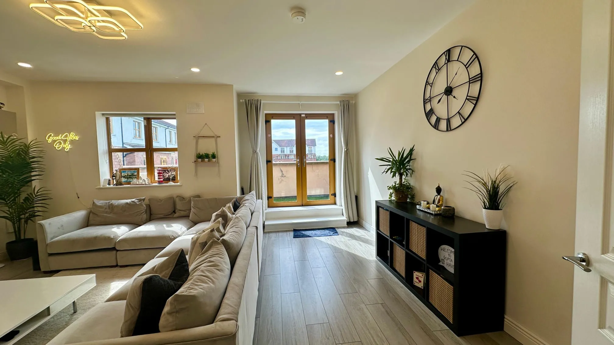 Living room with an L-shaped sofa, low coffee table, potted plants, a large wall clock, and double glass doors leading to a small balcony.