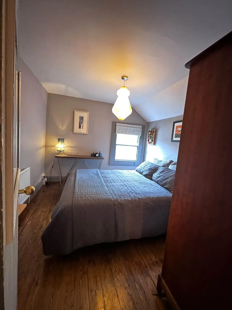 Bedroom with a large bed, window with a blind, bedside table and lamp, a small desk against the far wall, and a wardrobe at the right edge.
