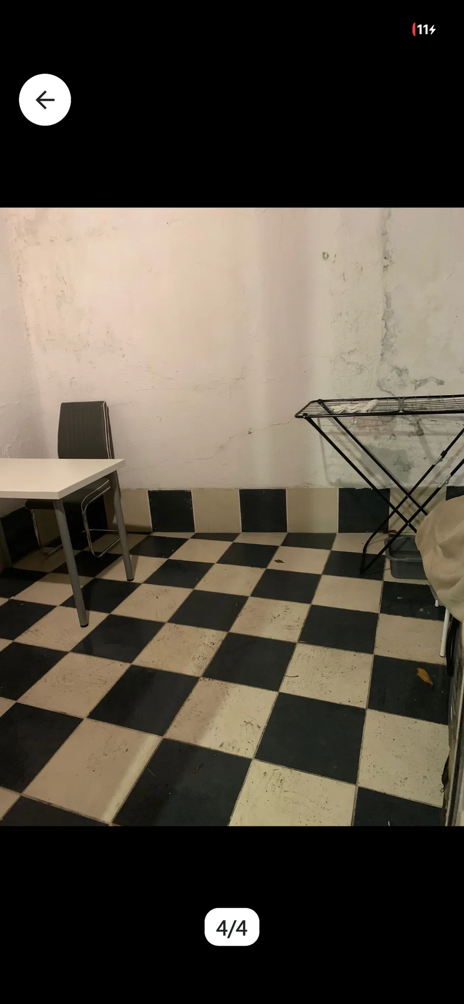 Room with black-and-white checkered tile floor, a small table and chair at left, and a foldable drying rack against a white wall with visible cracks and stains.