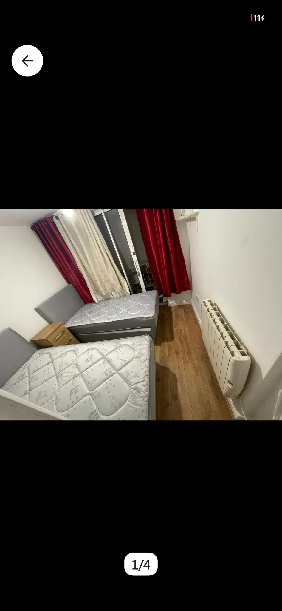 Small bedroom with two single beds, mattress tops visible, a wooden nightstand between beds, red and cream curtains covering a sliding glass door, and a wall-mounted radiator.