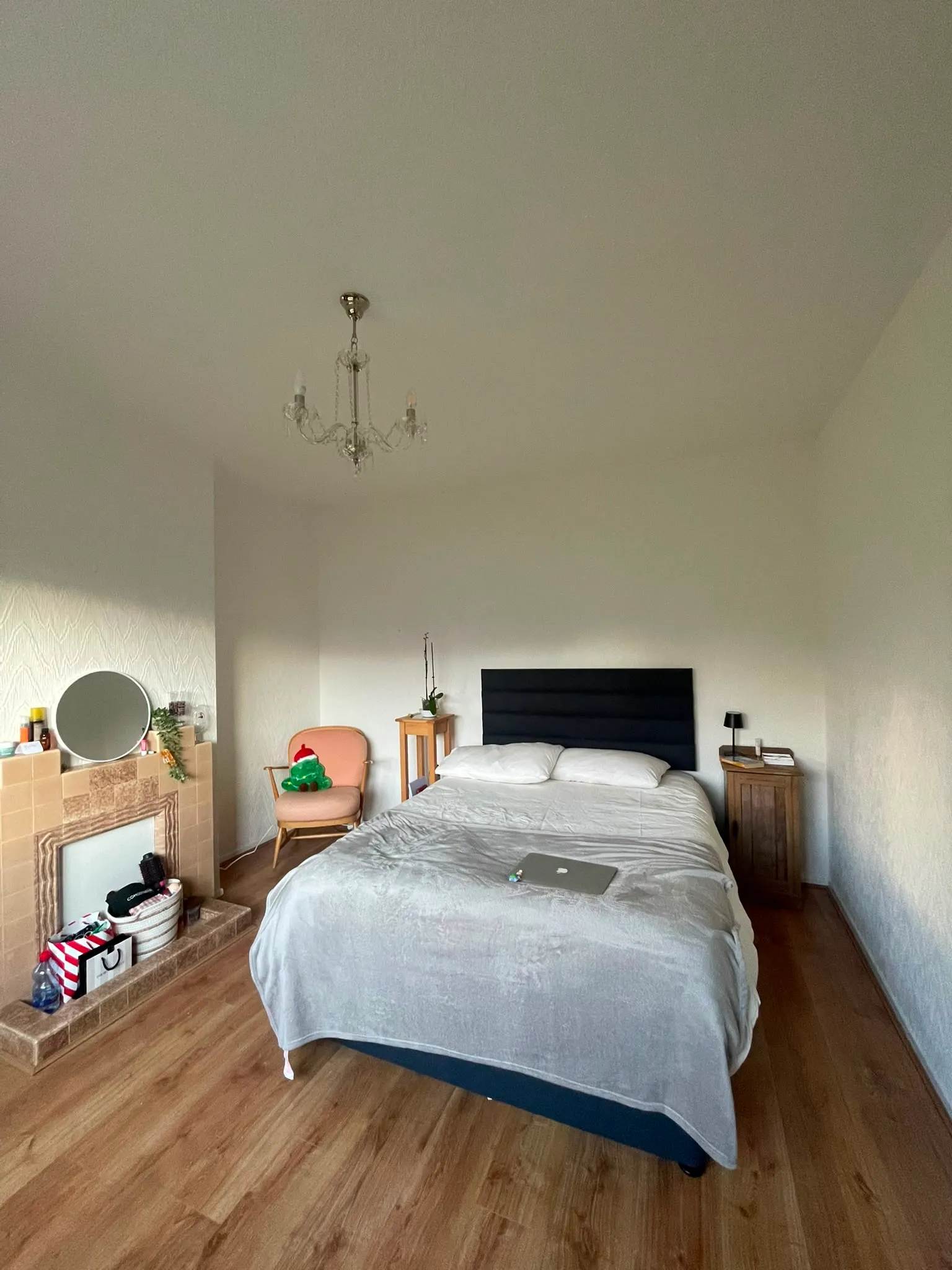 Bedroom with a double bed, dark headboard, bedside tables with a lamp, a chandelier, an armchair, and a fireplace with a round mirror.