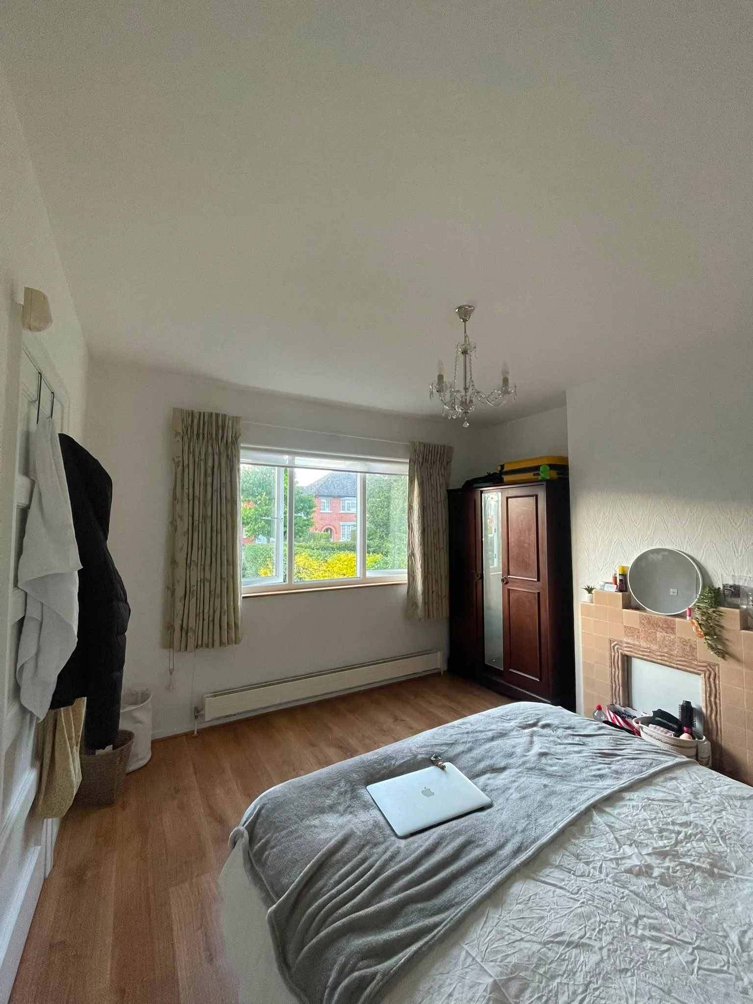 Bedroom with a bed in the foreground, window with curtains, wooden wardrobe, fireplace mantel, and a ceiling chandelier.