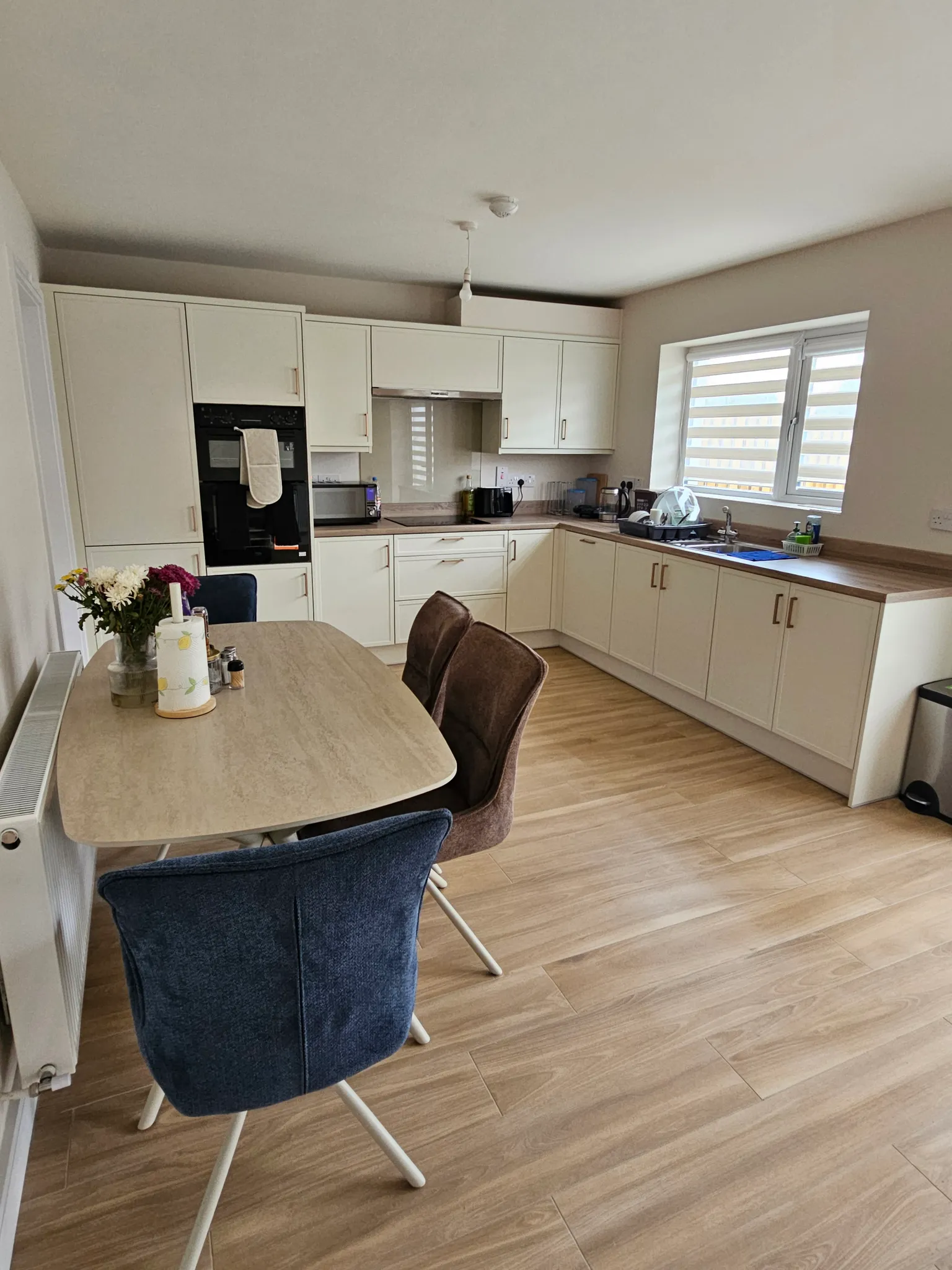 Kitchen with L-shaped white cabinets, countertop, built-in oven, sink beneath a window, dining table with chairs and flowers.