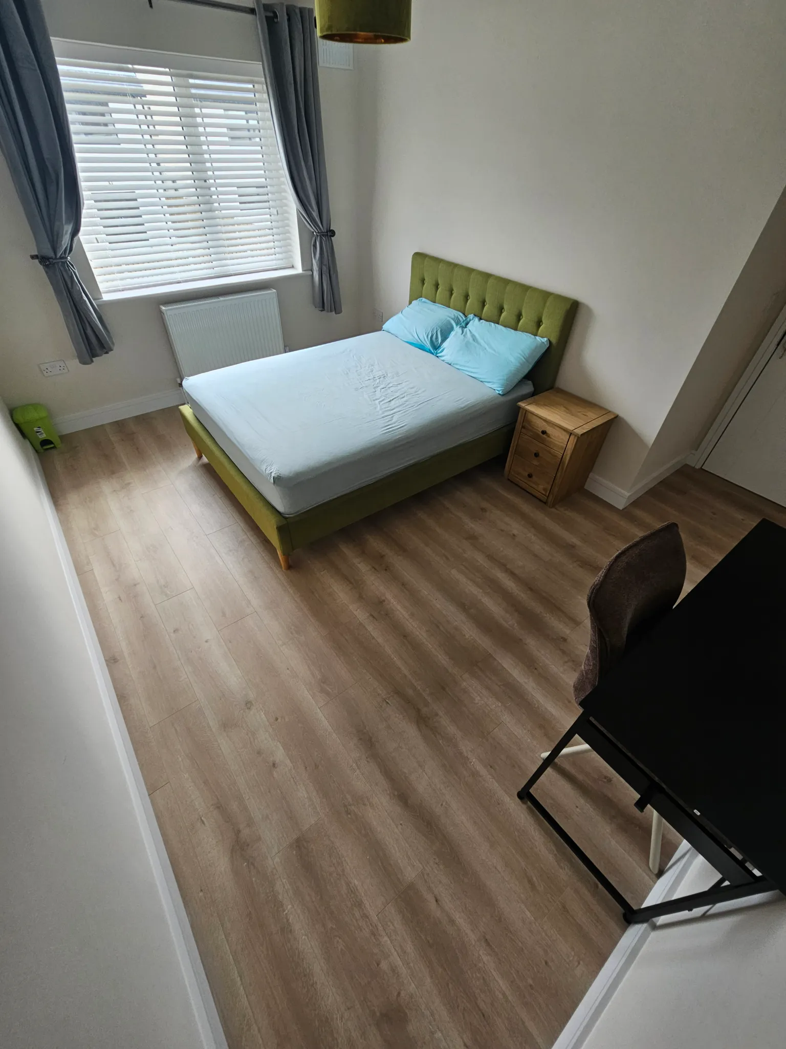 Bedroom with a double bed with green tufted headboard, two pillows, wooden bedside drawer, window with blinds and grey curtains, desk and chair, and wood-effect flooring.